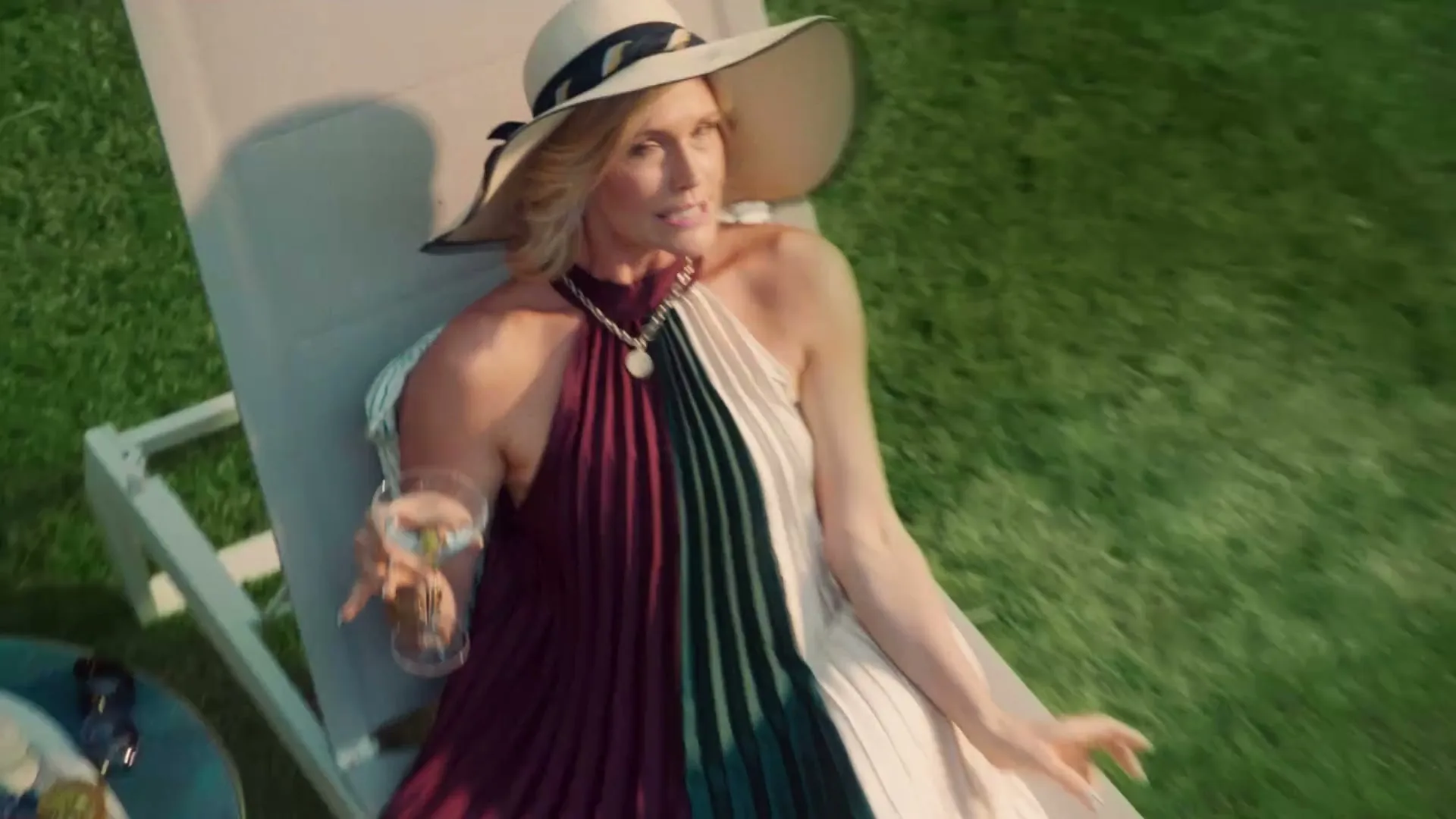 A woman in a wide-brimmed hat and a colorful dress reclines on a lounge chair, holding a cocktail and smiling at the camera in a sunny backyard.
