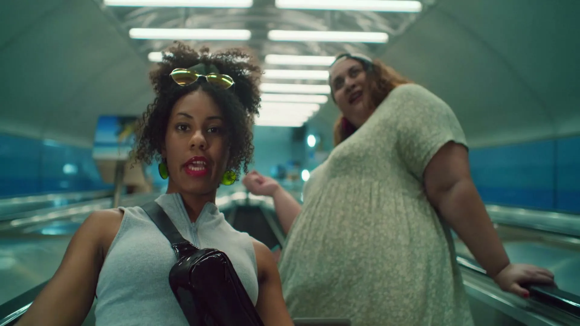 Two women, one with curly hair and sunglasses, the other in a floral dress, ride an escalator in an airport or subway station, looking excited.