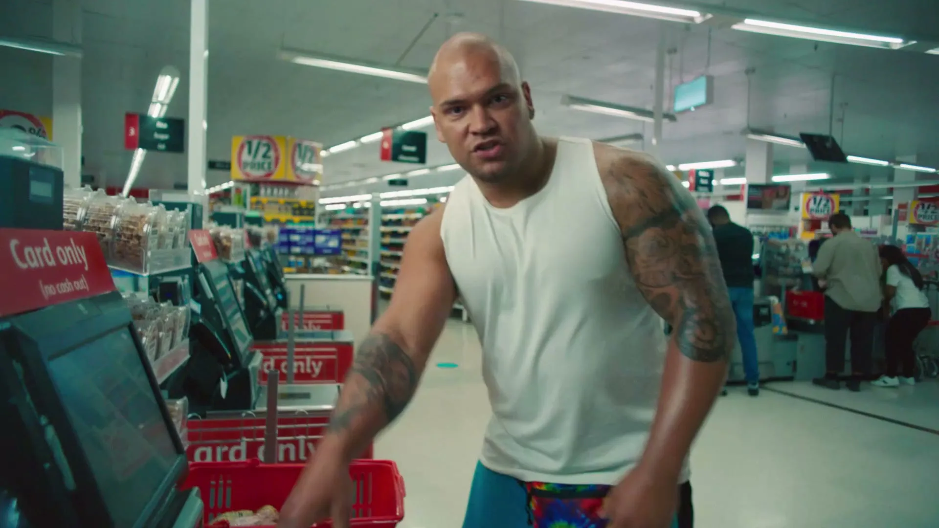 A bald, muscular man with tattoos on his arms, wearing a white tank top and a colorful fanny pack, looks directly at the camera while standing at a self-checkout in a brightly lit supermarket.