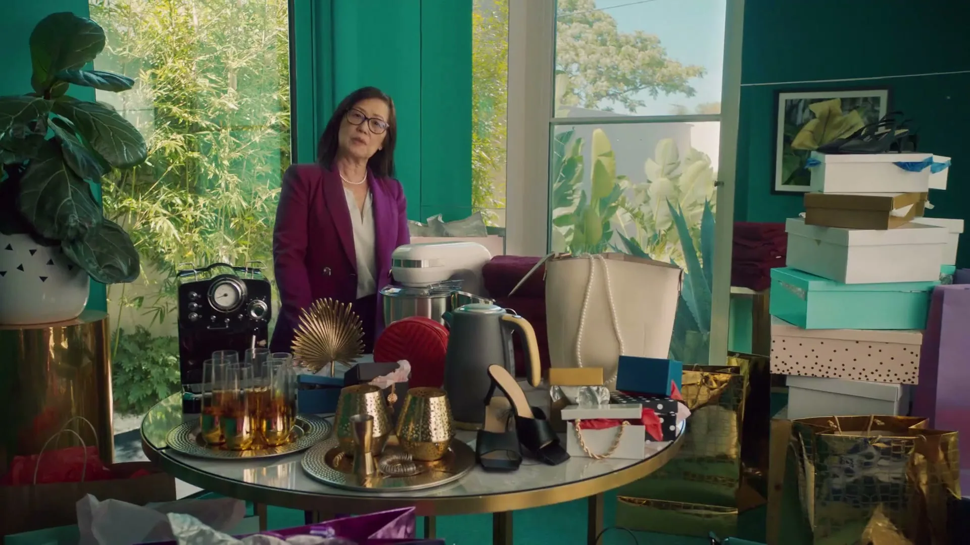 A woman in a purple blazer stands behind a table laden with gifts, looking directly at the camera in a brightly lit room.