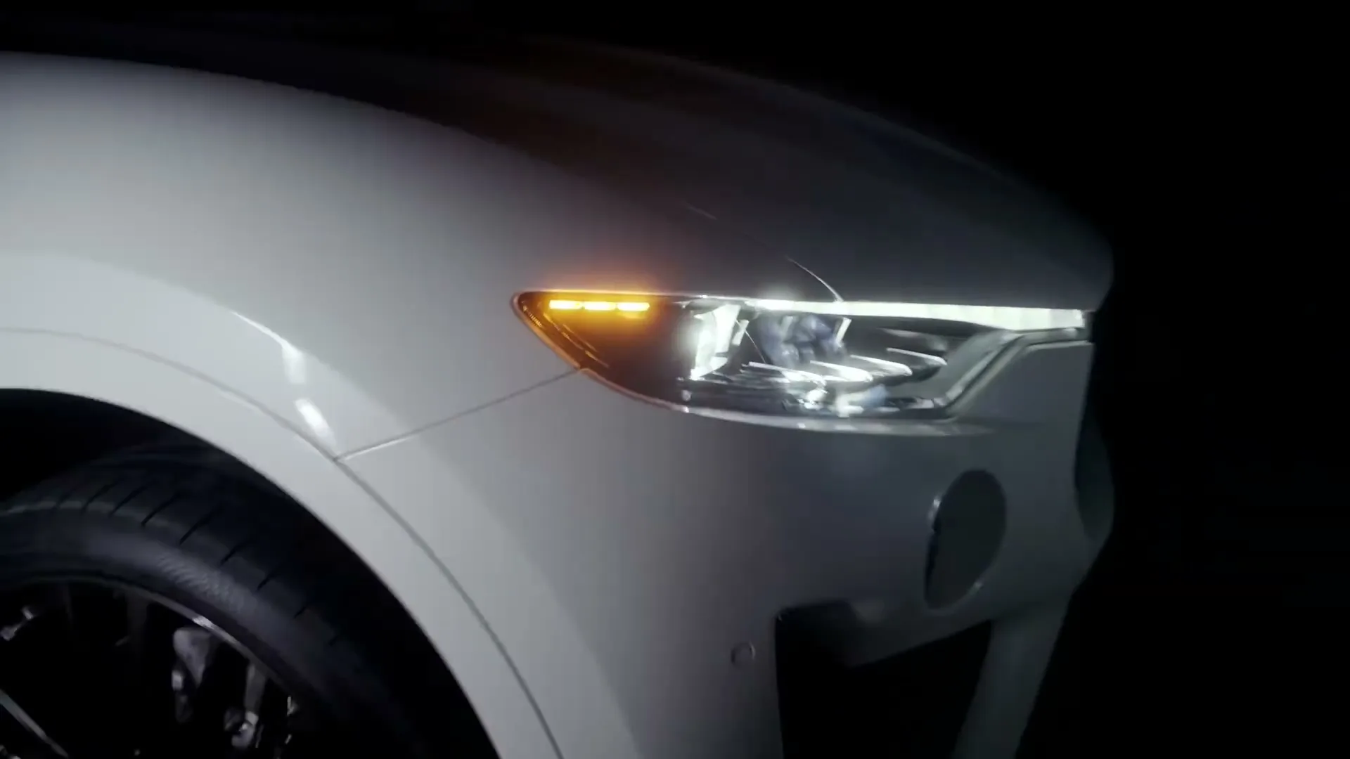A white Maserati car's front headlight and turn signal illuminate in a dark studio, highlighting its sleek design.