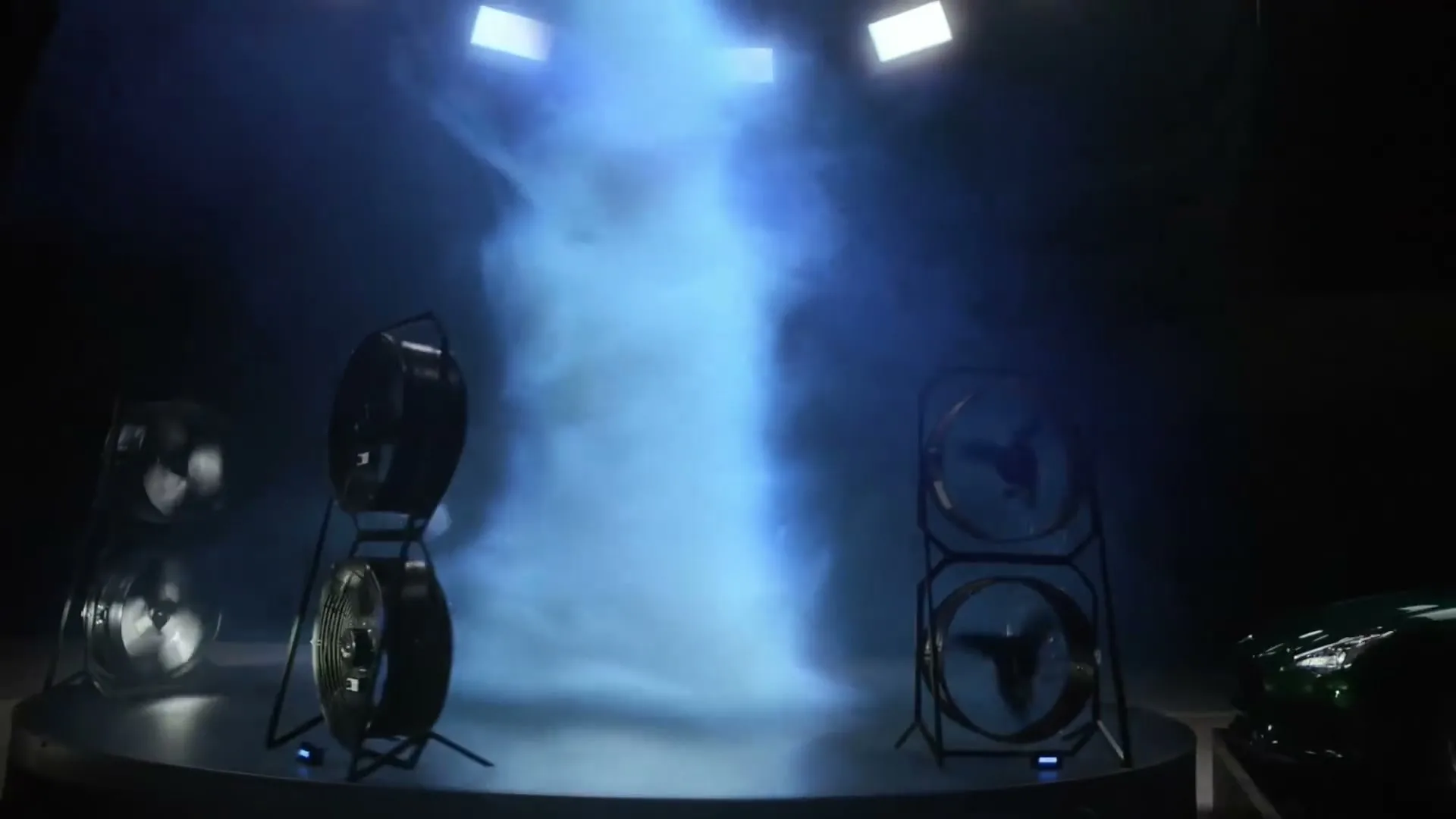 A red and a green Maserati car are parked in a dark studio with large fans blowing smoke upwards, illuminated by blue light.