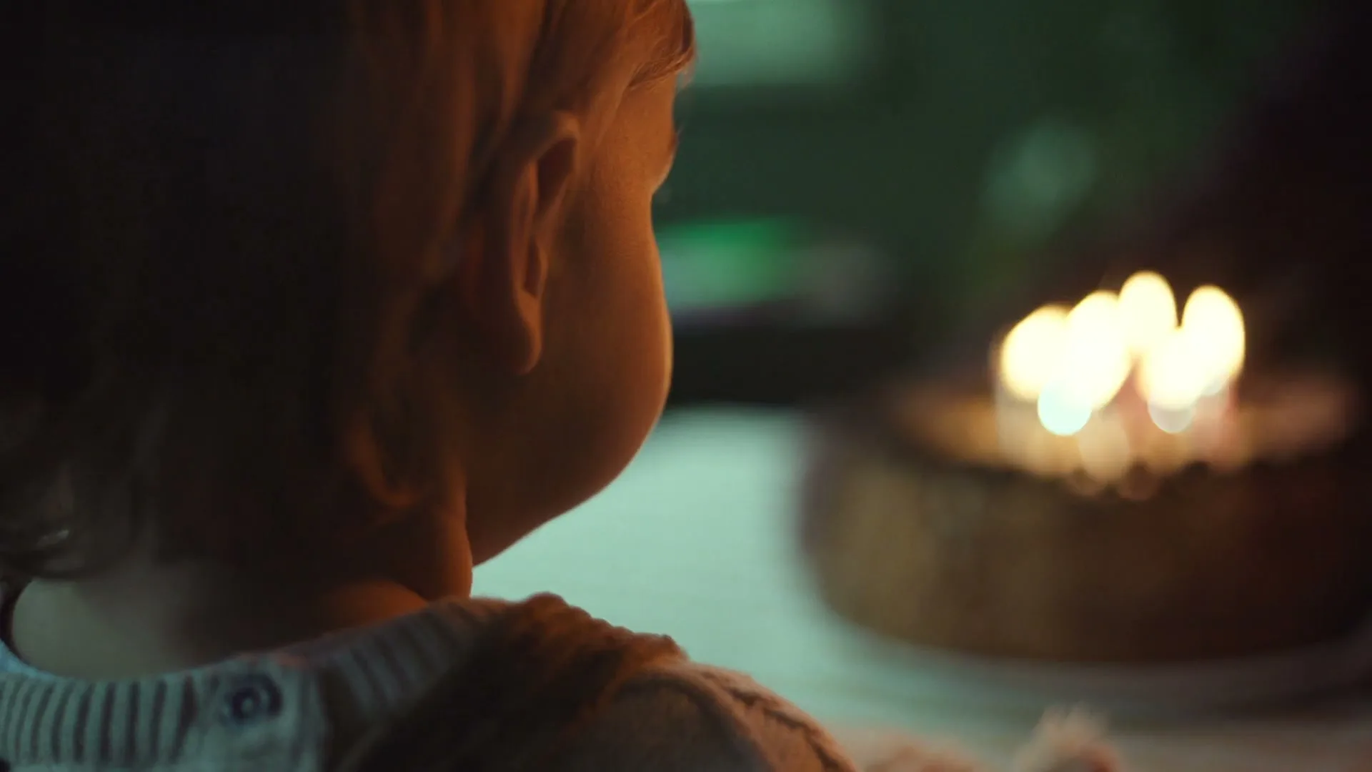 A baby with blonde hair, wearing a knitted sweater, looks at a birthday cake with lit candles in a dimly lit room.