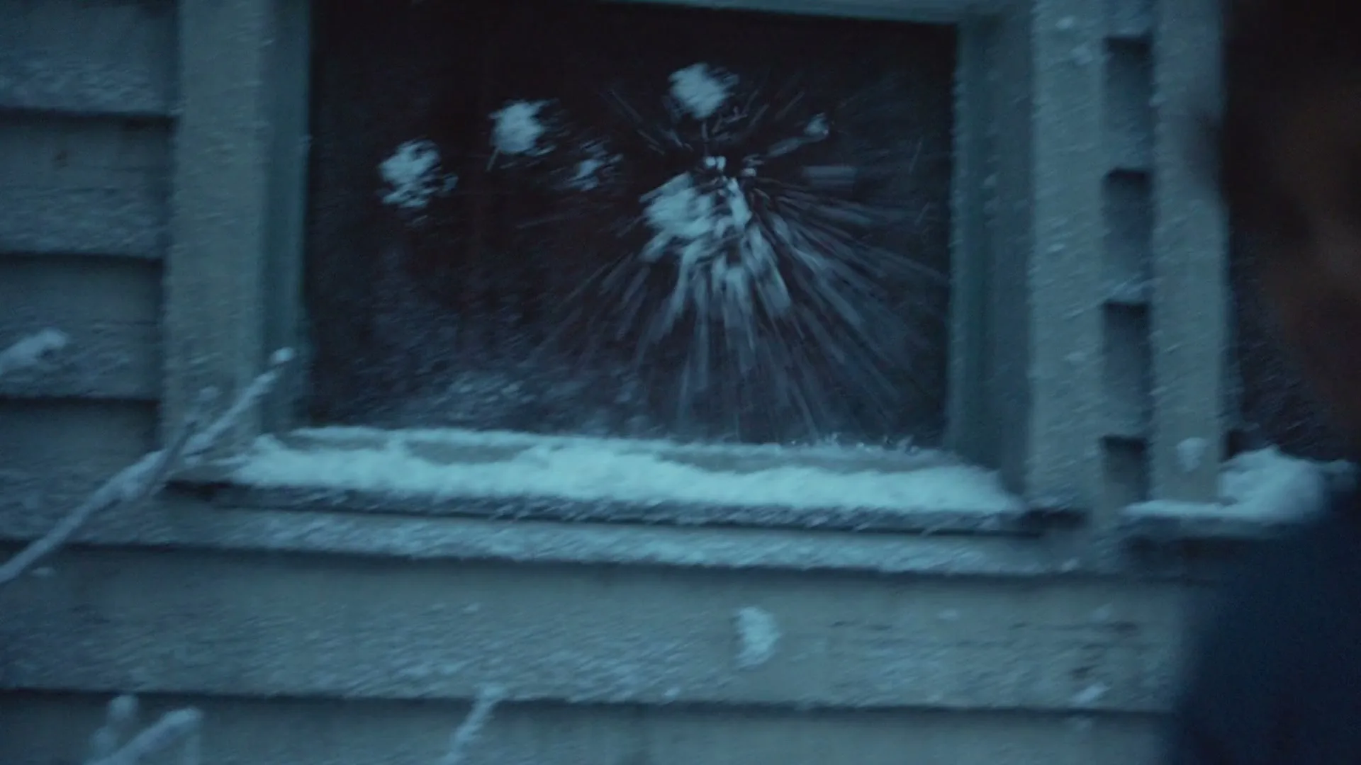A child in a blue coat throws a snowball at a snow-covered window, causing snow to splatter on the glass.