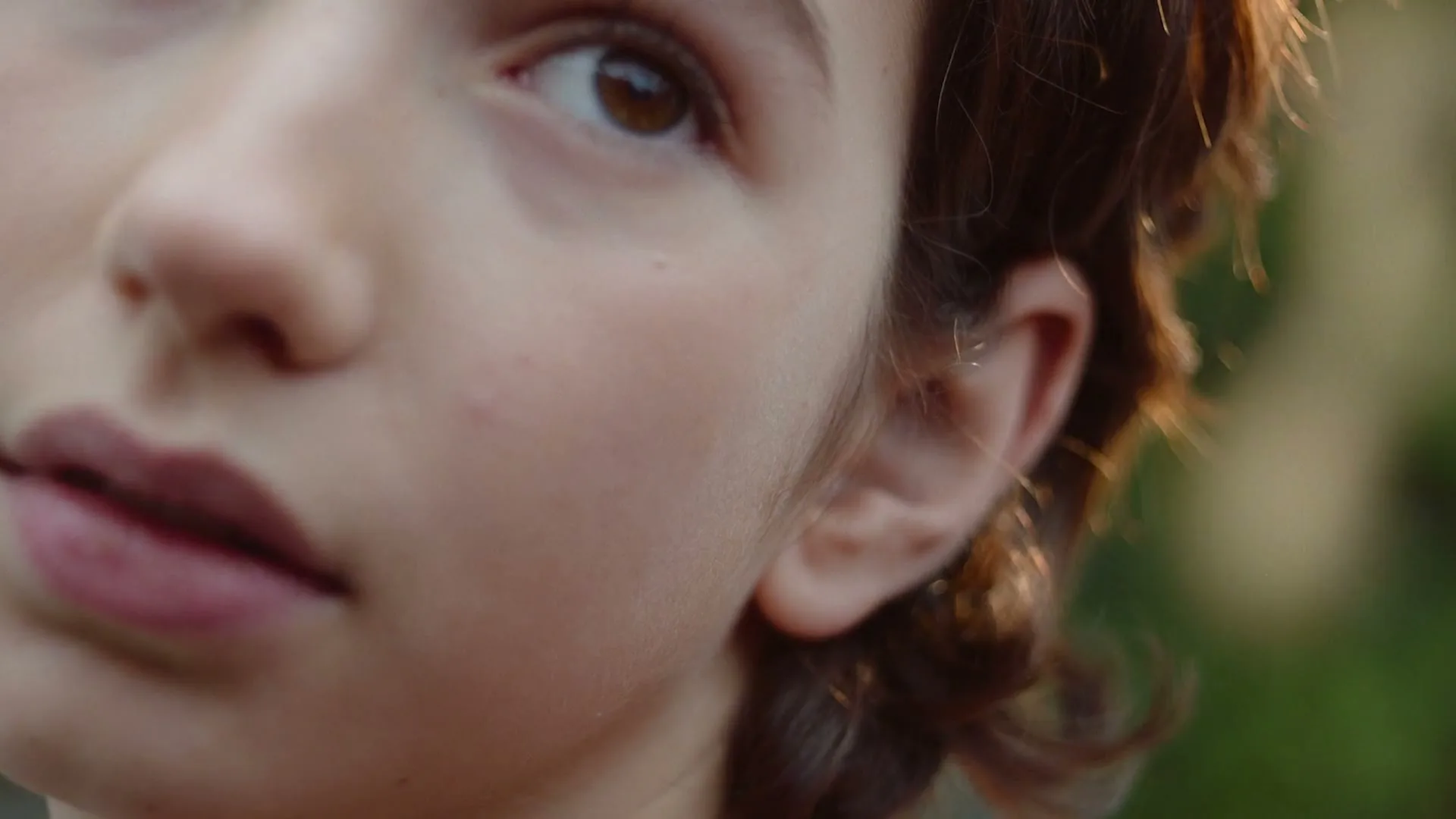A young person's face is shown in a close-up, with their ear and part of their eye visible, against a blurred green background.