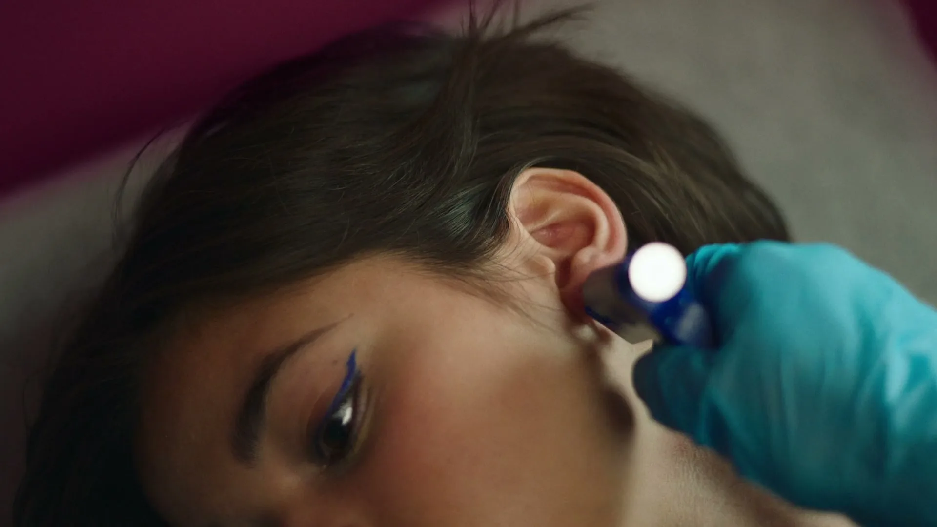 A person with dark hair and blue eyeliner lies down as a gloved hand holds a small device near their ear, possibly for an ear piercing.