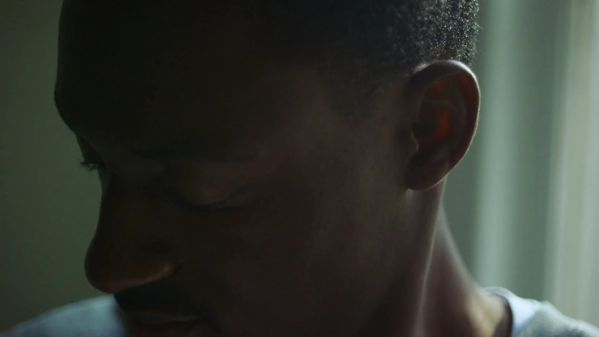 A young Black man with short hair and a white t-shirt looks down, his face partially shadowed by natural light from a window.