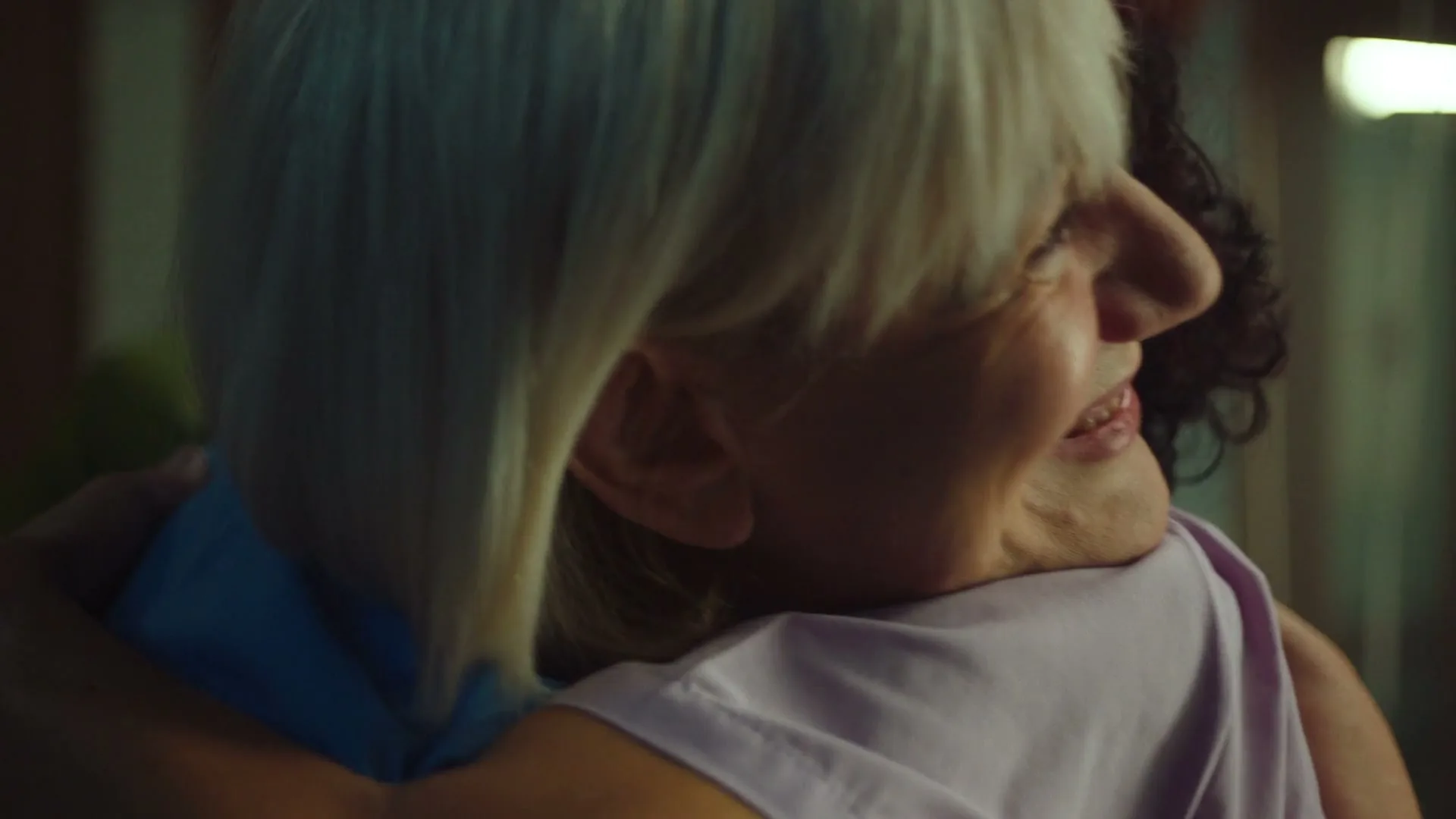 A blonde woman with a hearing aid smiles while hugging a person with dark curly hair, seen from the side in a warm interior.