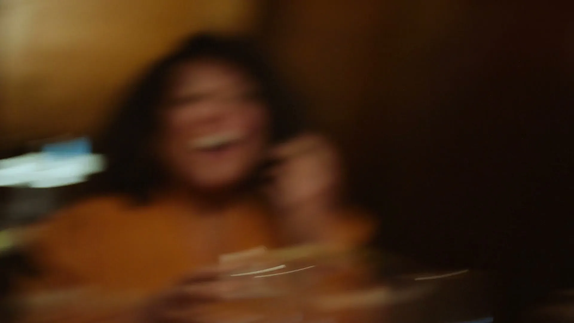 A blurry shot of a woman with dark hair, wearing an orange top, laughing and holding a glass, in a dimly lit interior.