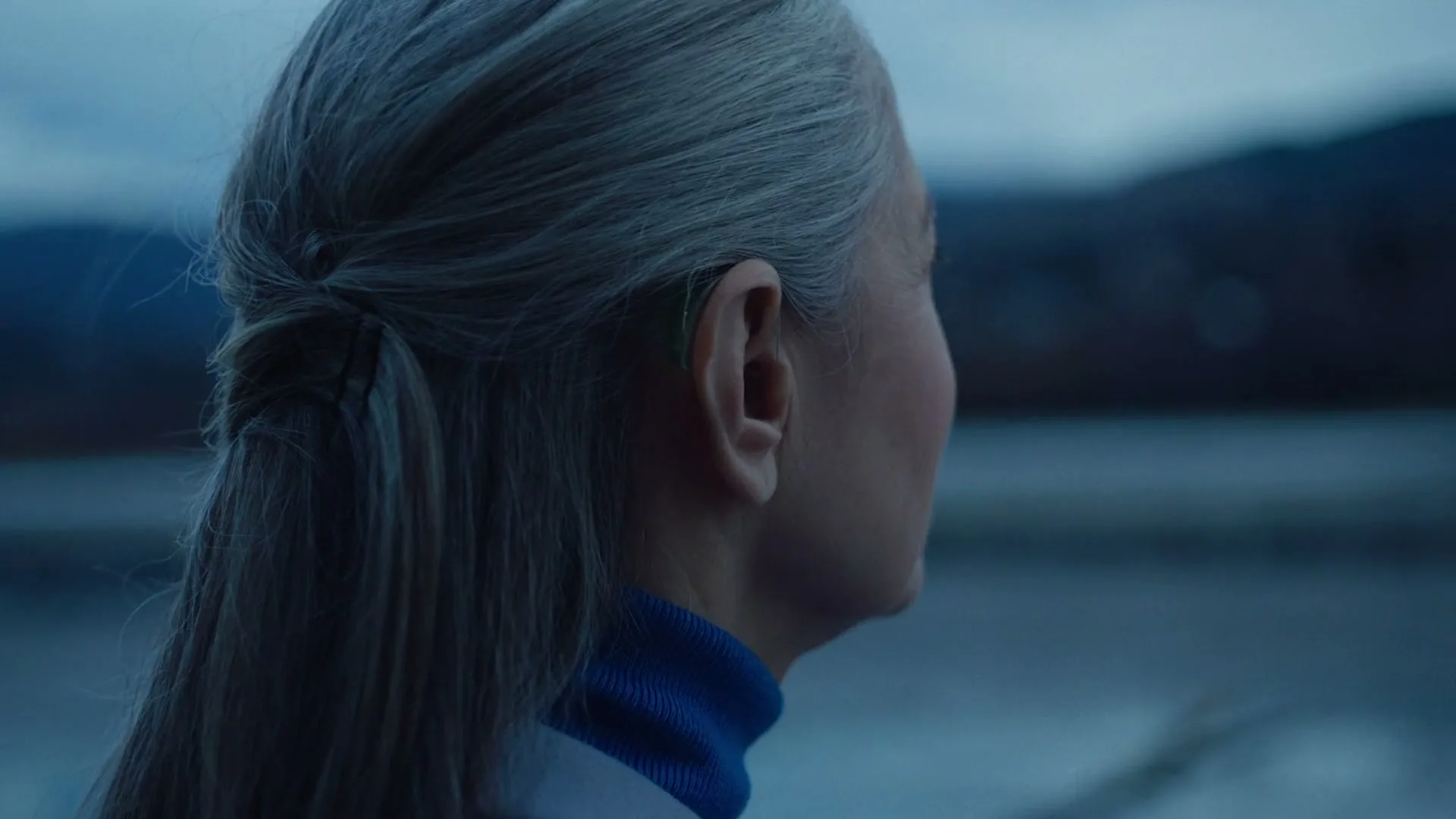 A senior woman with gray hair and a blue turtleneck adjusts her hearing aid while looking out at a lake with mountains in the background.