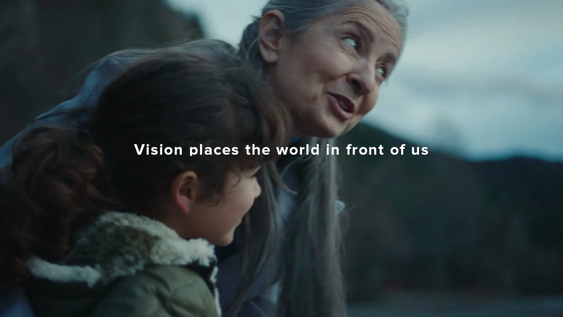 A grandmother with long grey hair and a young girl with curly brown hair look out at a lake with mountains in the background.