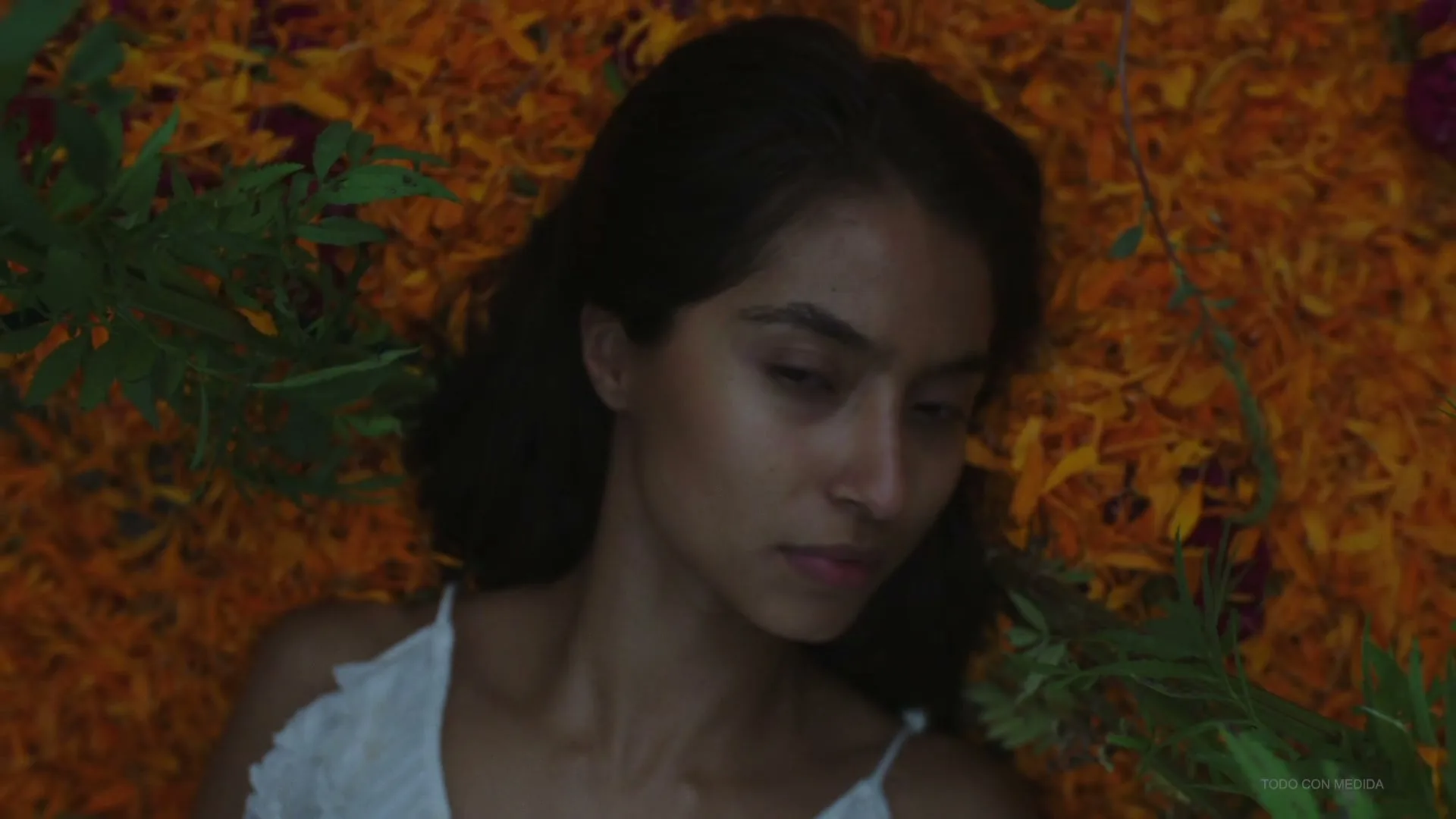 A young woman with dark hair and a white top lies on a bed of orange and yellow flower petals, slowly opening her eyes.