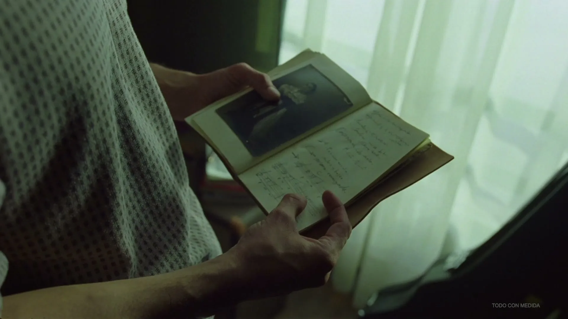 A person holds an old, open book with a faded photograph and handwritten notes, standing near a window with sheer curtains.