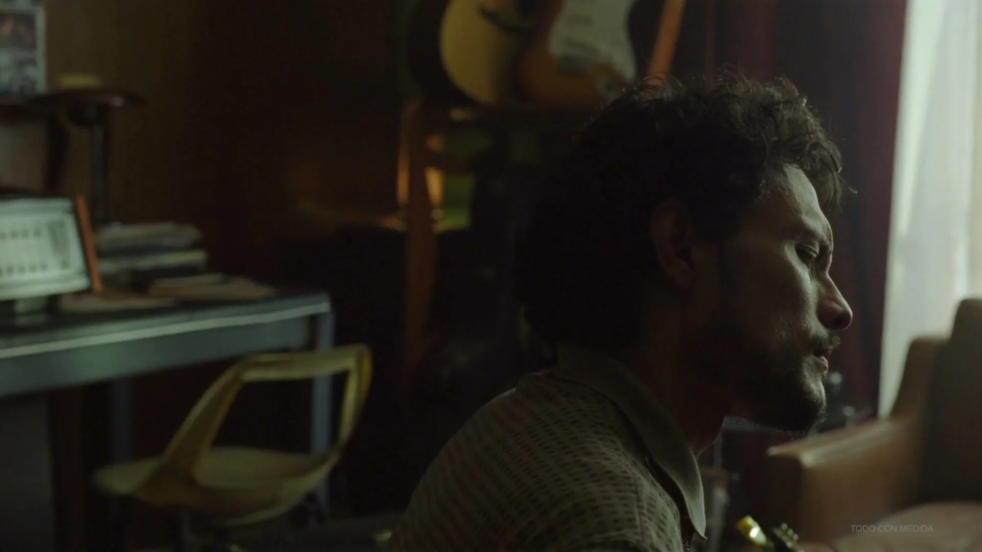 A man with dark curly hair and a beard looks off-screen to the right, sitting in a dimly lit room with musical instruments in the background.