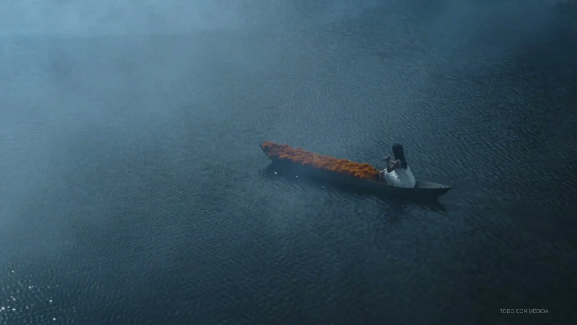 A person in a white dress sits in a small boat filled with orange flowers, drifting across a dark, misty lake.