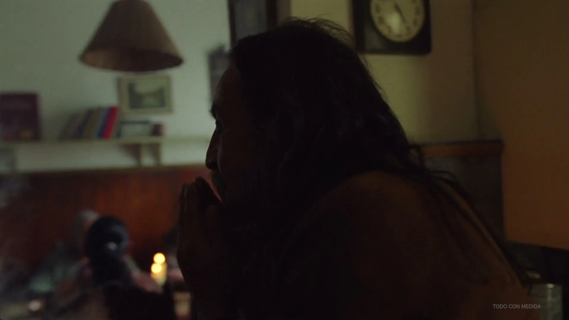 A man with long dark hair sits in profile at a table, dimly lit by candles, with other people in the background.
