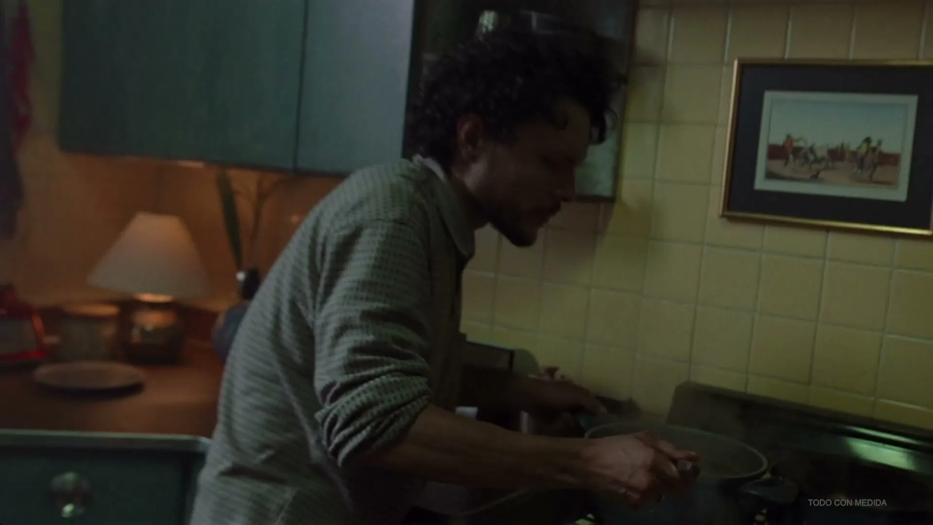A man with curly hair and a beard cooks on a gas stove in a dimly lit kitchen, stirring a pot with a spoon.