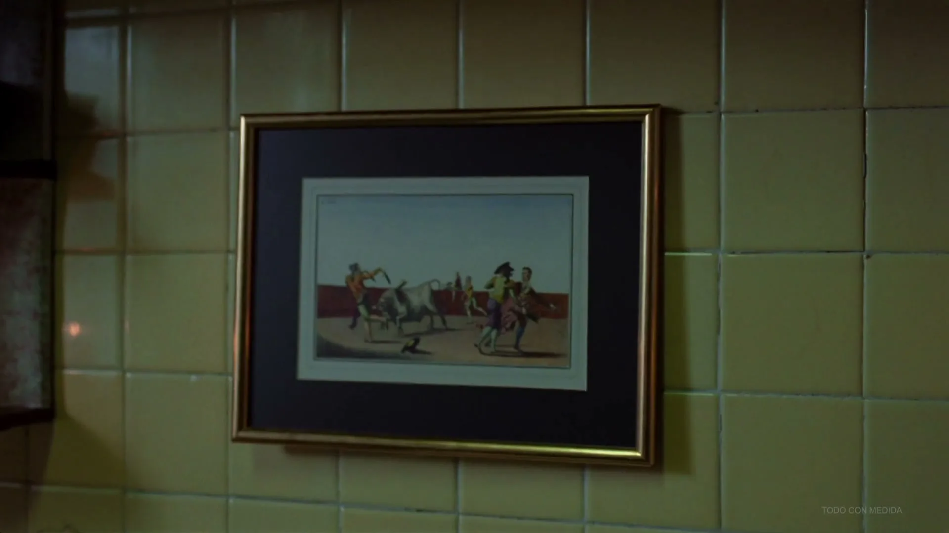 A framed painting depicting a bullfight hangs on a tiled wall in a dimly lit interior setting.