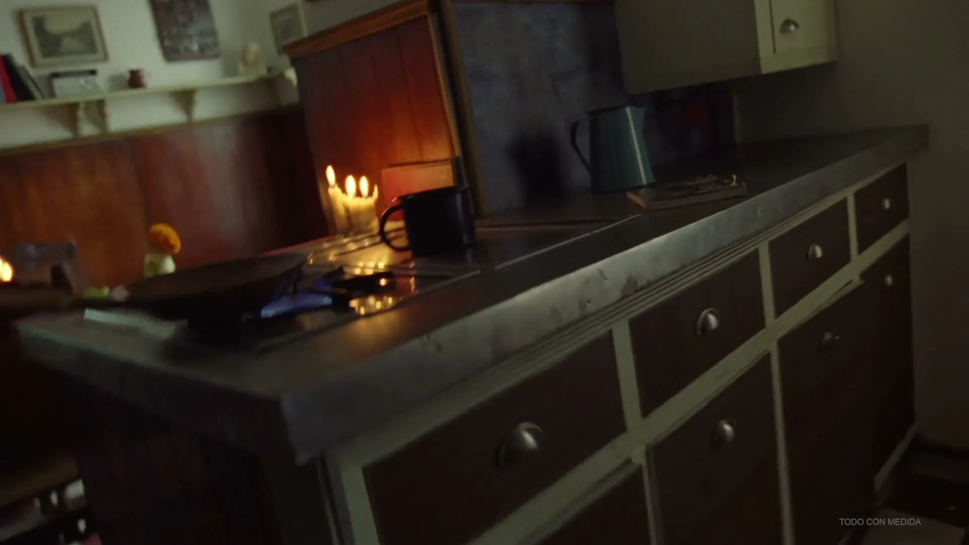 An older man with long gray hair and a beard looks off-screen, then the camera pans to a kitchen counter with lit candles and a stove.