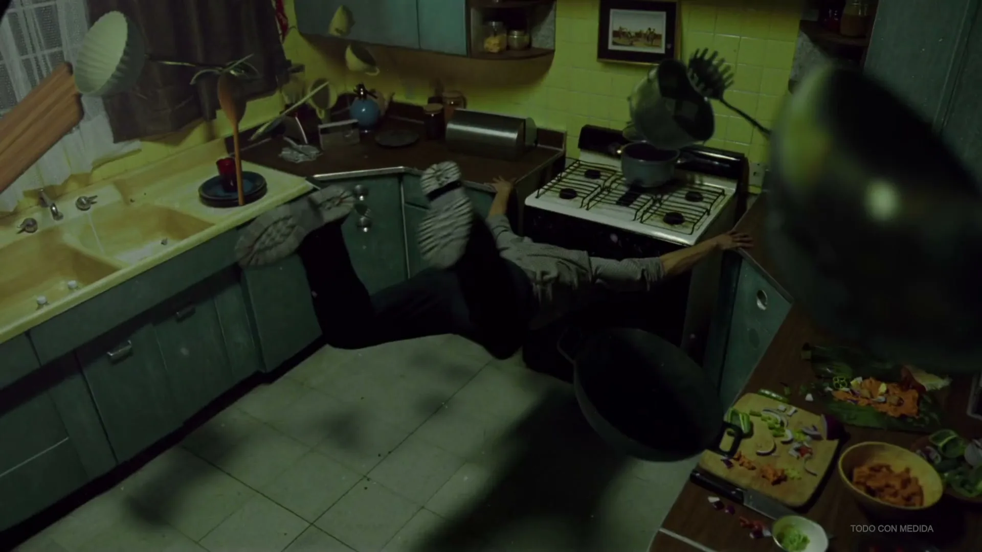 A person in dark clothes and a grey shirt is seen floating horizontally in a kitchen, with kitchenware floating around them.