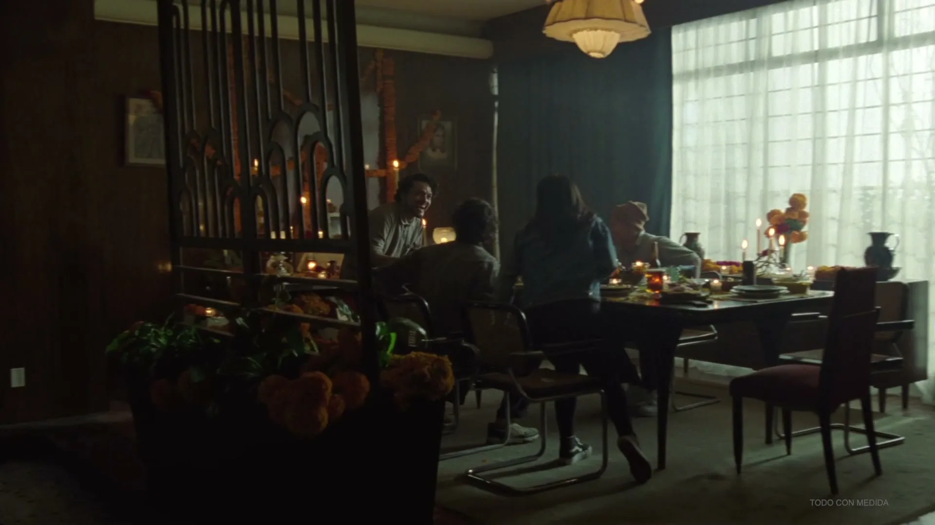 A group of friends gathers around a dining table, adorned with candles and marigolds, in a dimly lit room with a large window.