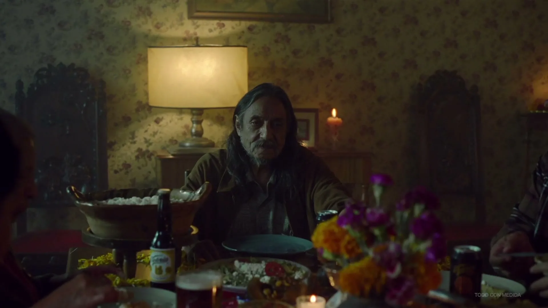 A man with long dark hair and a beard sits at a dinner table, looking forward with a neutral expression, surrounded by food and drinks in a dimly lit room.