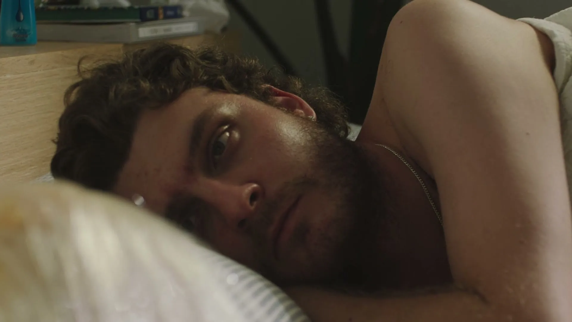 A man with curly hair and a beard lies in bed, looking directly at the camera with a somber expression in a dimly lit room.
