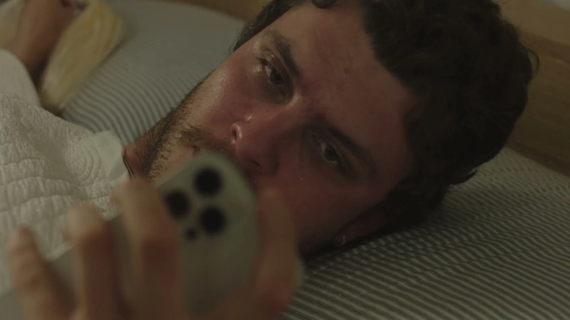 A young man with dark hair and a beard lies in bed, looking at his phone with a slightly confused expression, as a blonde woman's hair is visible next to him.