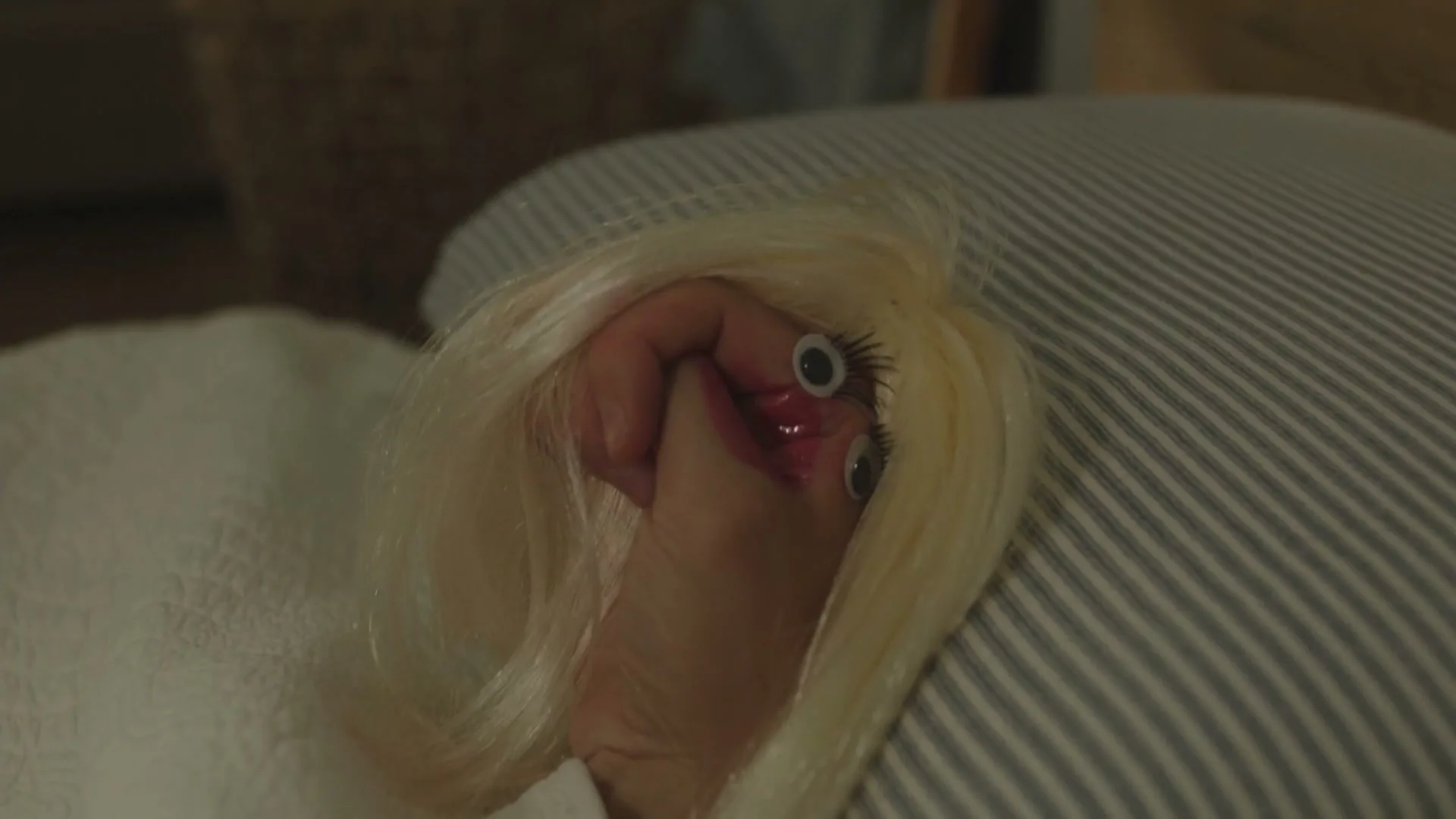 A hand puppet with blonde hair and googly eyes lies on a striped pillow, looking sad and dejected in a bedroom.