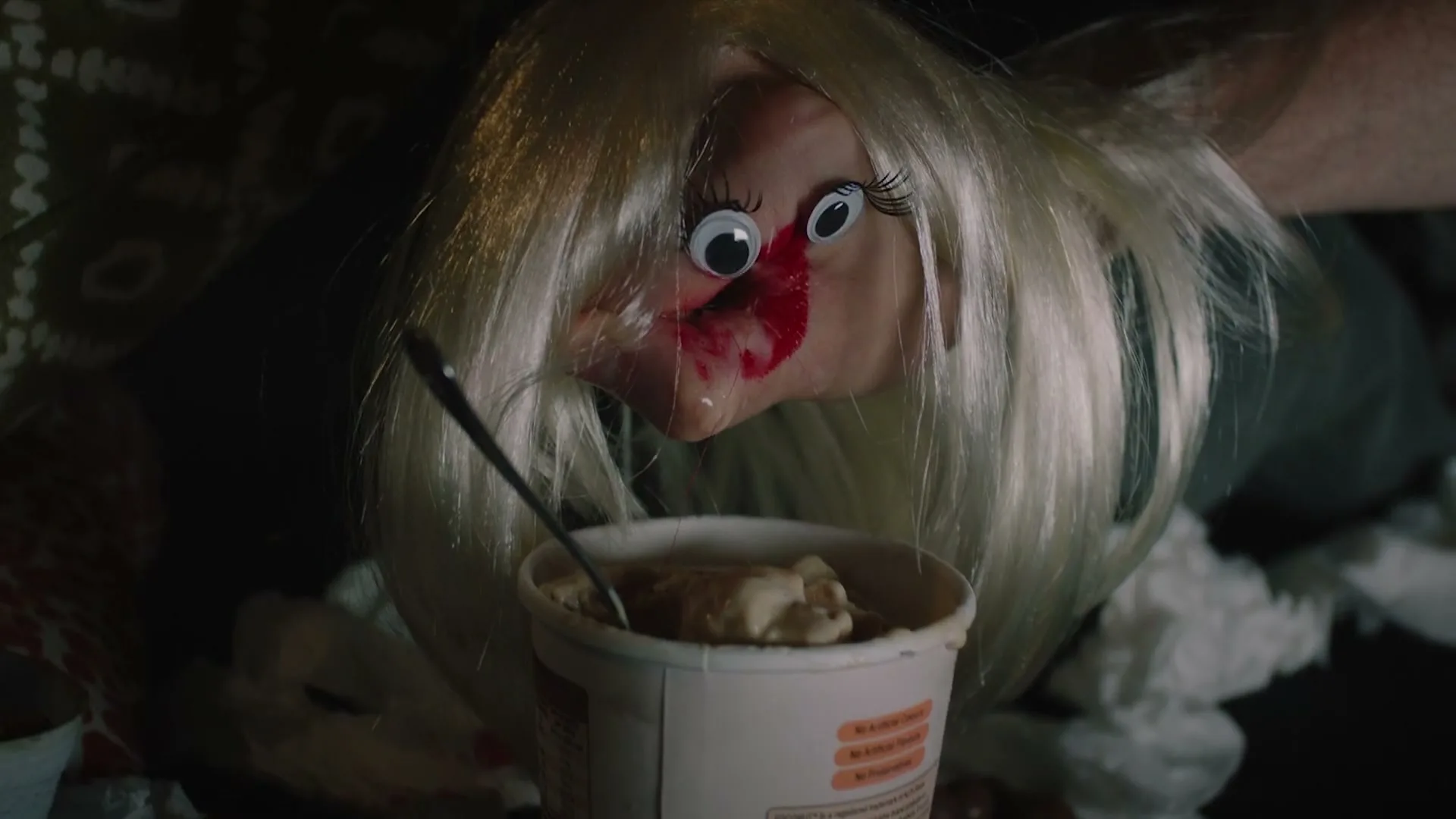 A person with a doll's head, blonde wig, and googly eyes eats ice cream from a tub with a spoon, surrounded by tissues in a dimly lit room.