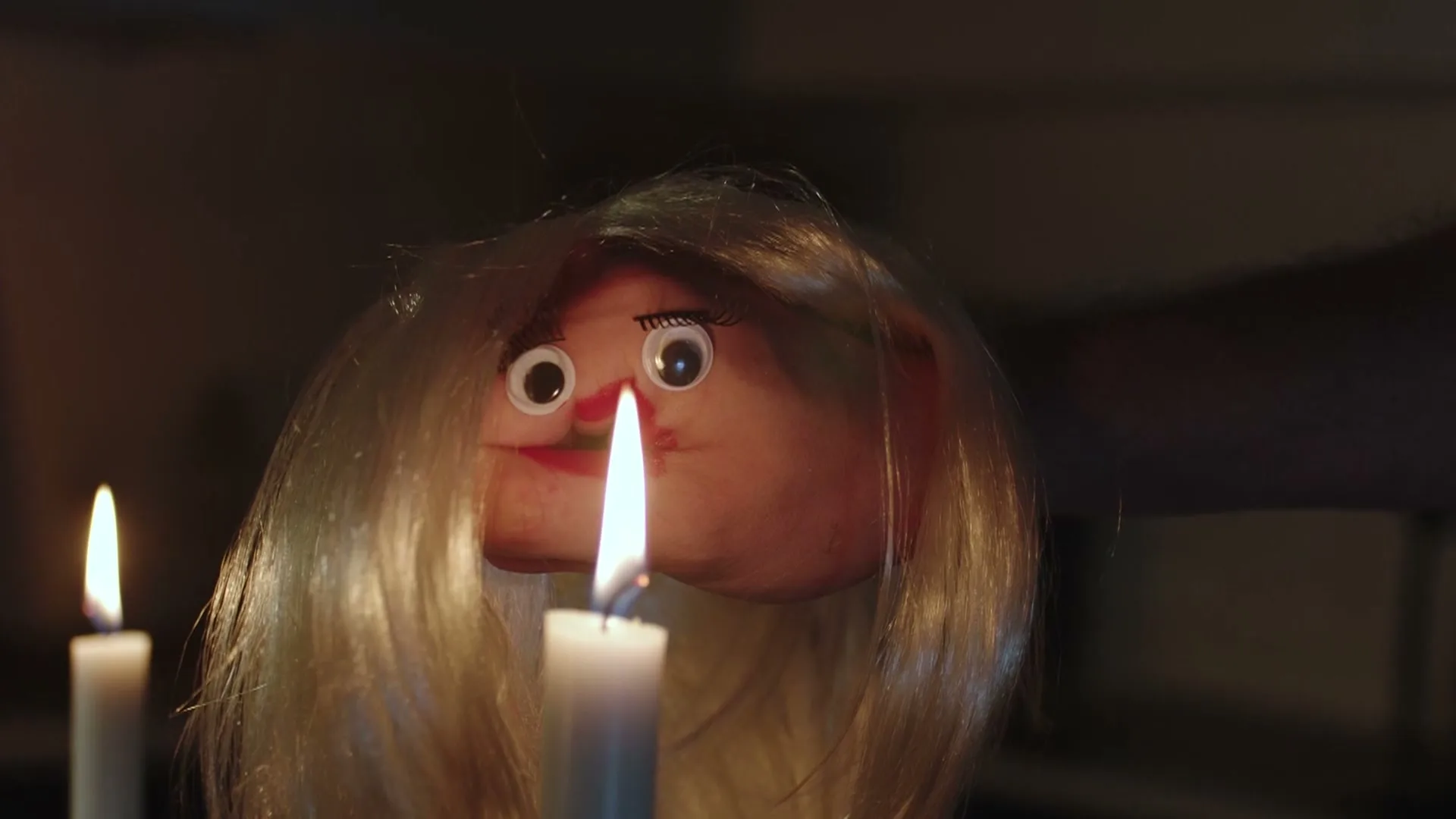 A doll's head with blonde hair and wide eyes is seen in a medium shot, with two lit candles in the foreground, creating a mysterious atmosphere.