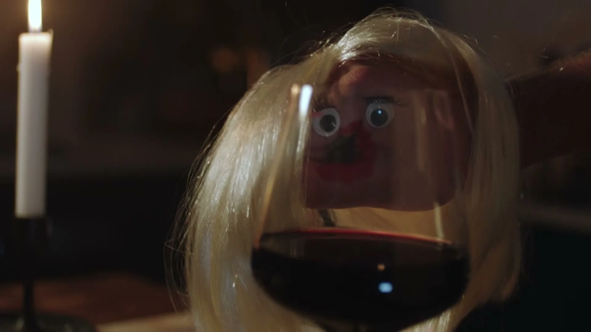 A hand puppet with blonde hair and googly eyes peers over a glass of red wine on a candlelit table in a dimly lit room.