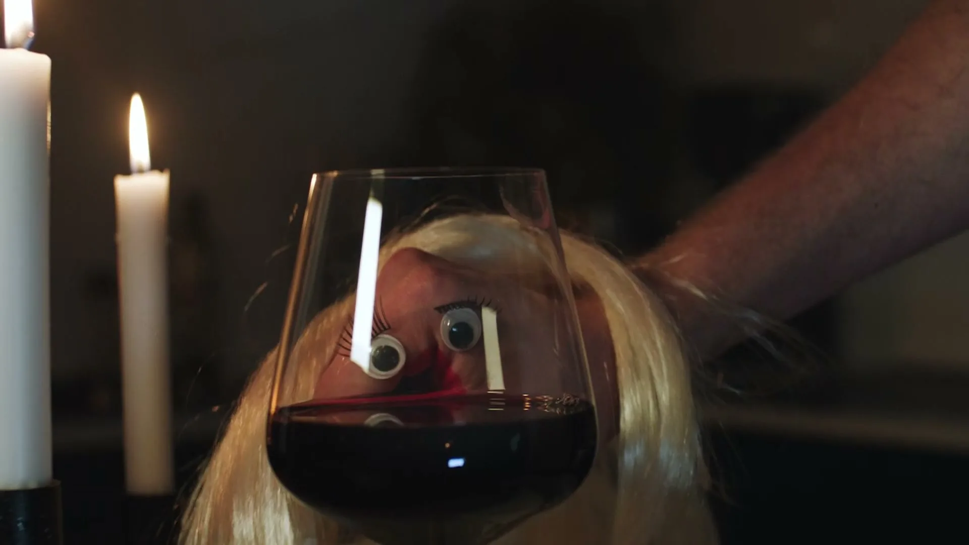 A person's hand with googly eyes and fake eyelashes attached to fingers, holding a wine glass with red wine, is seen in a dimly lit room with candles.