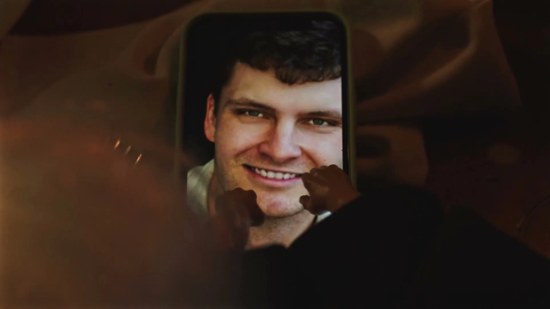 A man's smiling face is seen on a smartphone screen, with two small hands appearing to pinch his cheeks, in a dimly lit interior.