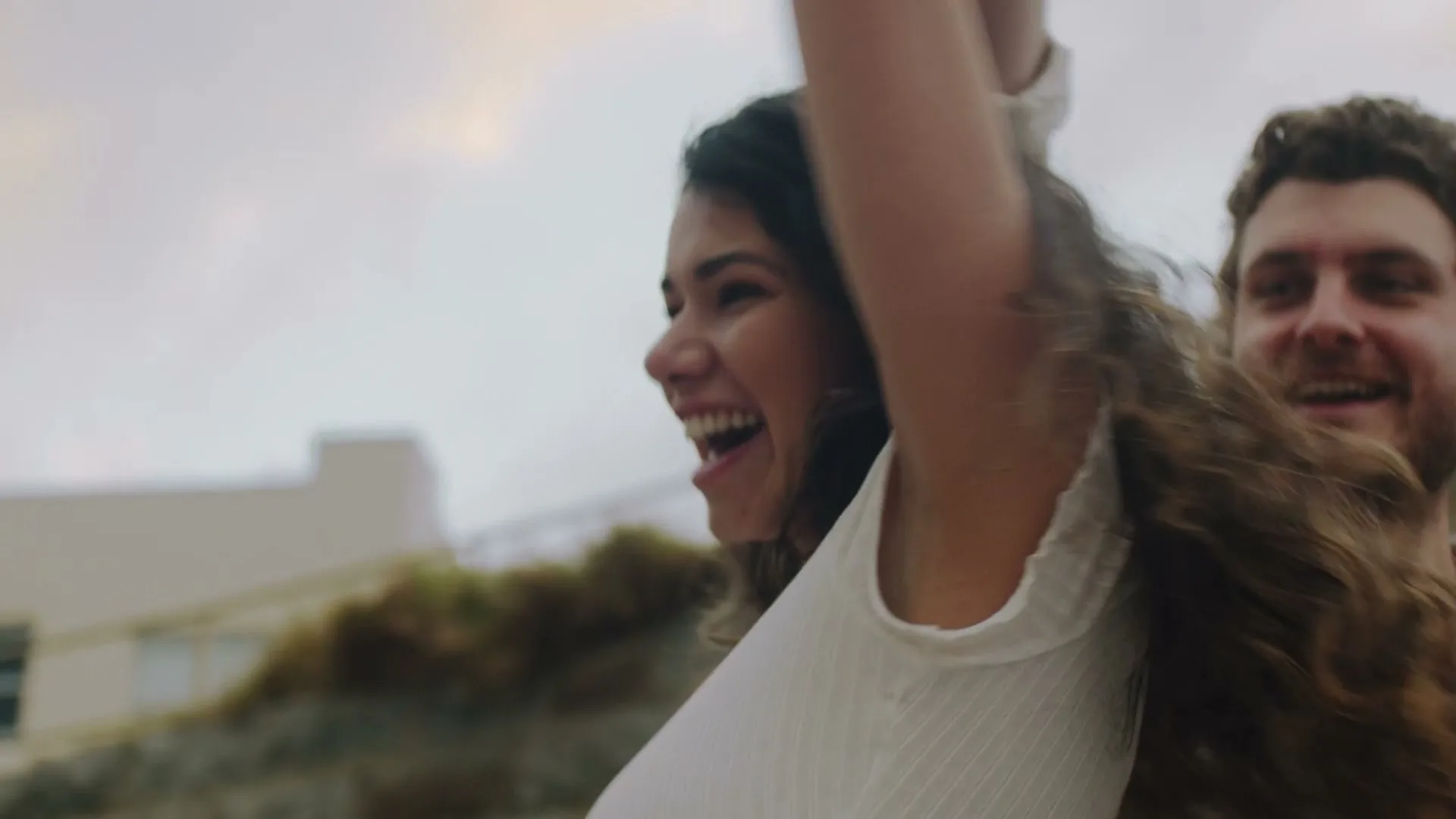 A young couple, a woman with long curly hair and a man with a beard, are joyfully running with their arms raised, laughing, outdoors near a building.
