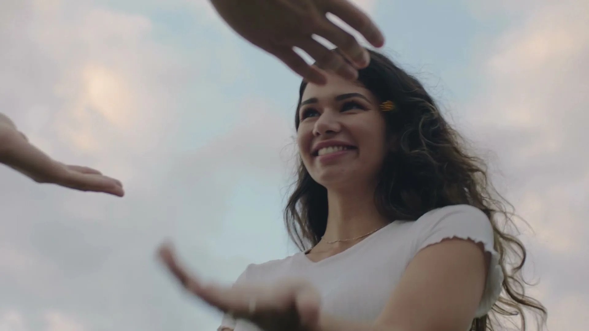 A young woman with long, dark, curly hair and a flower in her hair, wearing a white t-shirt, smiles and laughs as she reaches for hands against a cloudy sky.