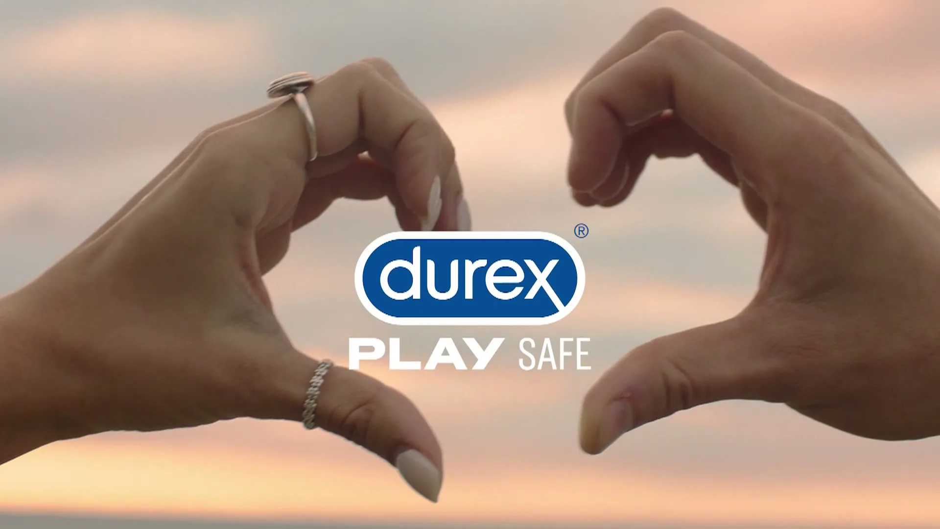 Two hands, one with a ring, form a heart shape against a soft, pastel-colored sky, with the Durex logo and "PLAY SAFE" text appearing in the center.