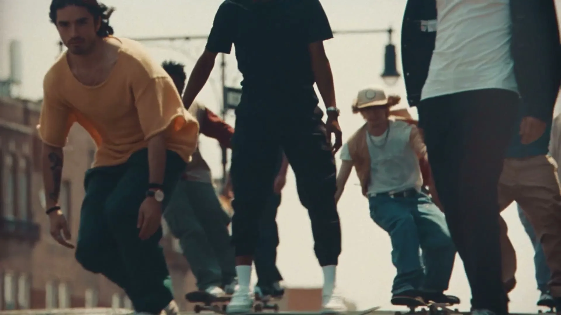 A diverse group of young adults skateboards down a city street on a bright day, with buildings faintly visible in the background.