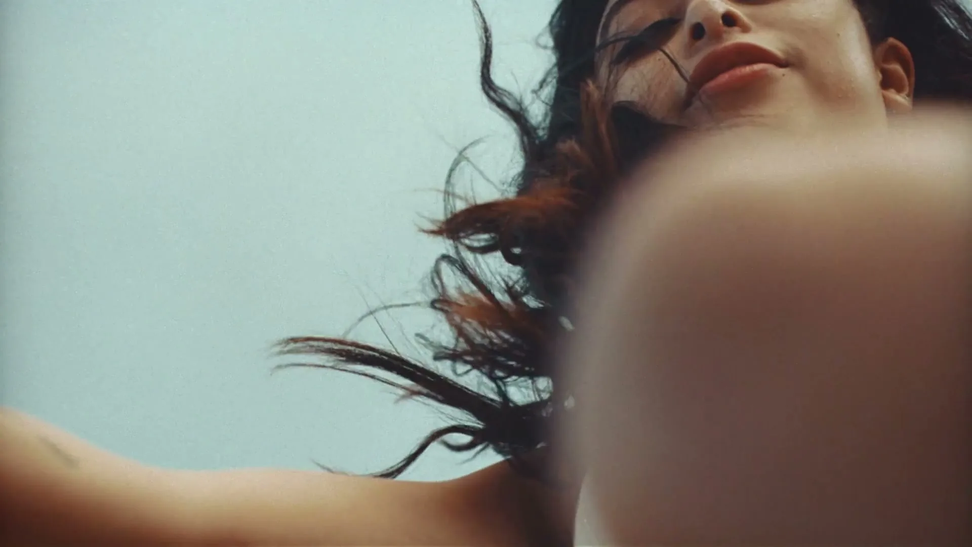 A young woman with dark hair and a white top looks down at the camera, her hair blowing in the wind against a light blue sky.