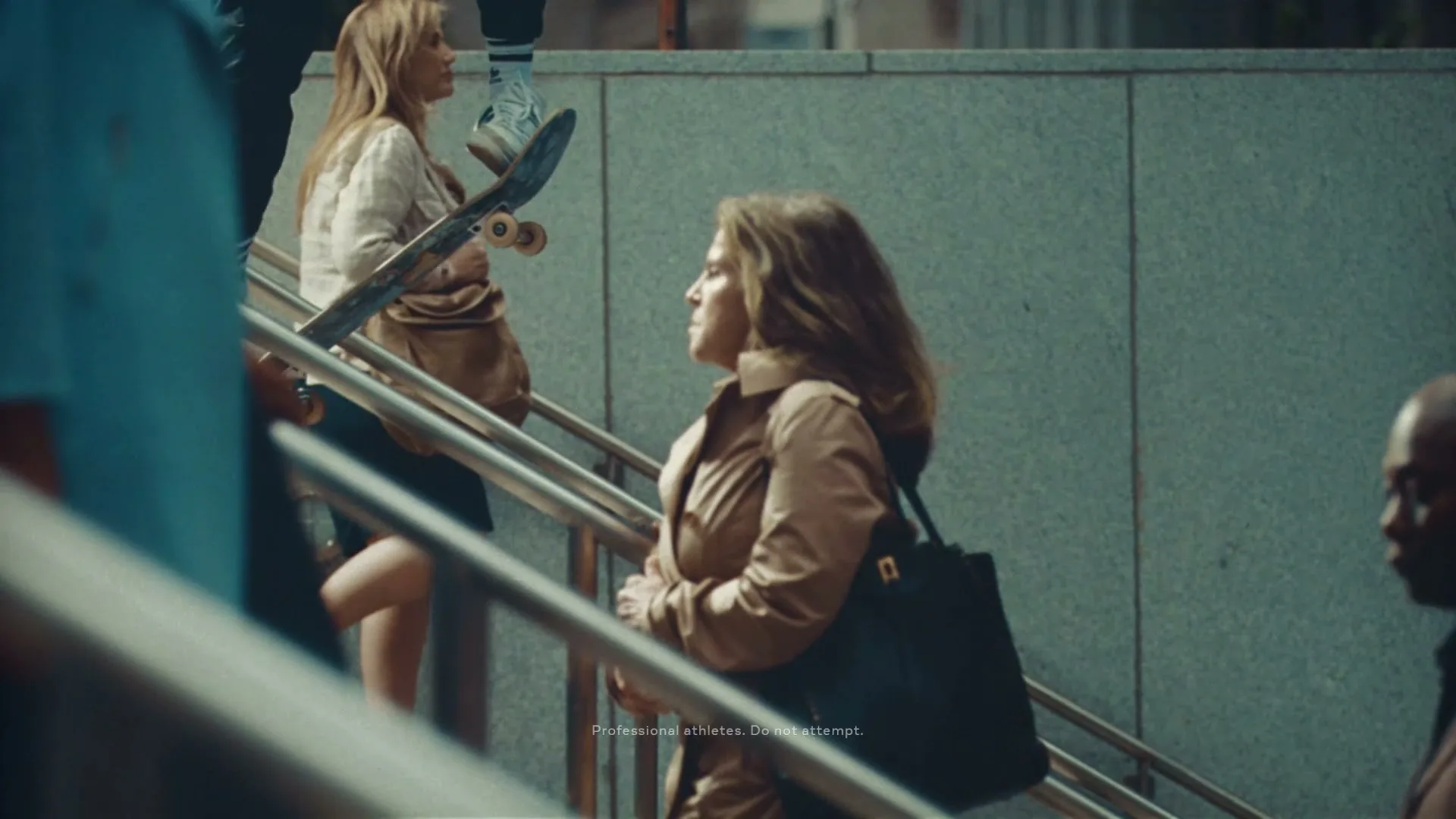 A young woman in a trench coat stands on an escalator, looking up, while a person on a skateboard ascends the adjacent escalator in a public transit station.