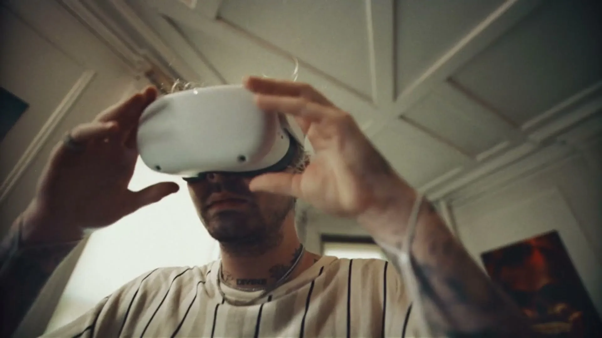 A man with tattoos and a striped shirt puts on a white VR headset in a room with a patterned ceiling.