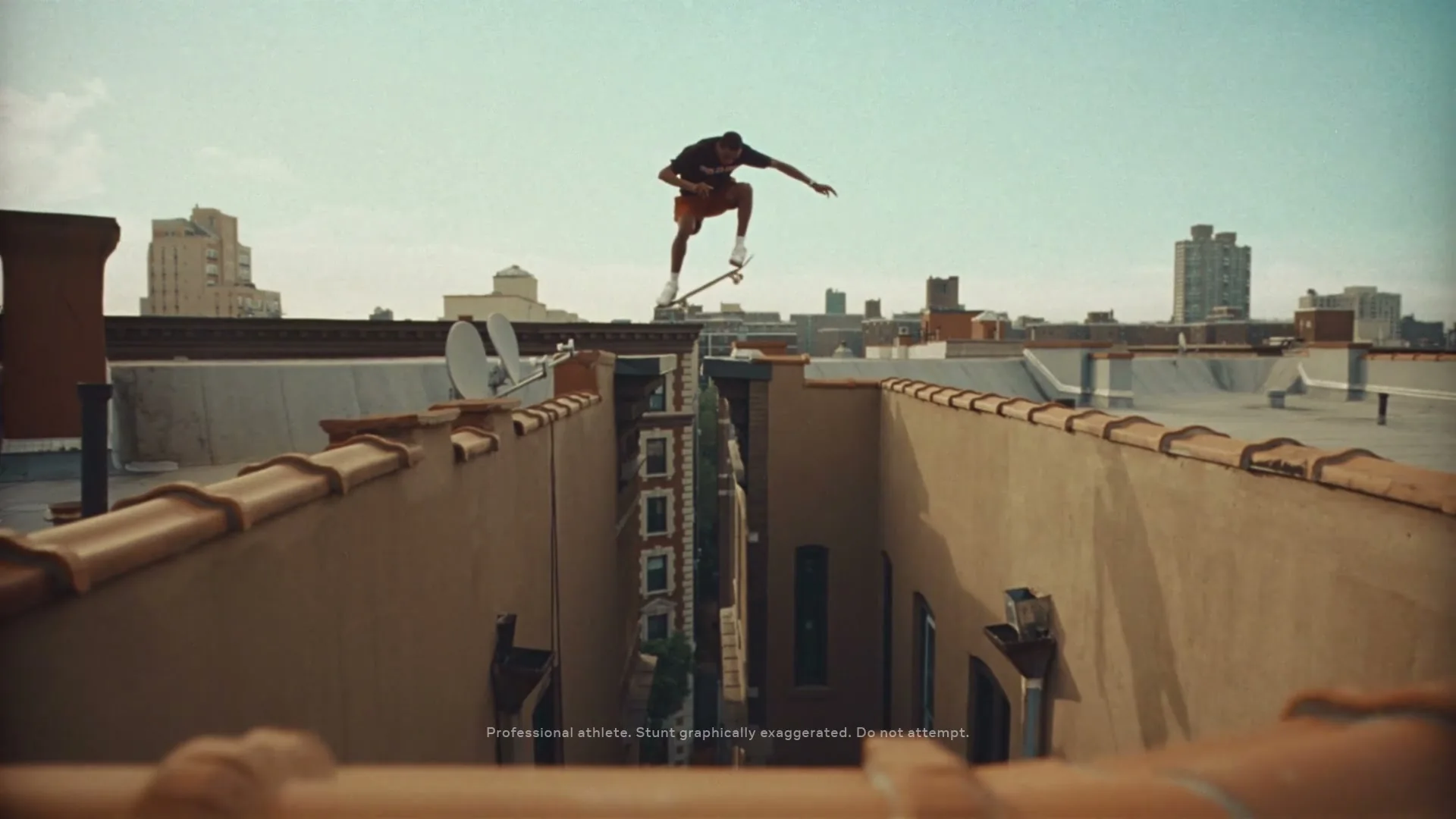 A male skateboarder in a black t-shirt and orange shorts performs an ollie over a gap between two buildings on a rooftop in a city.