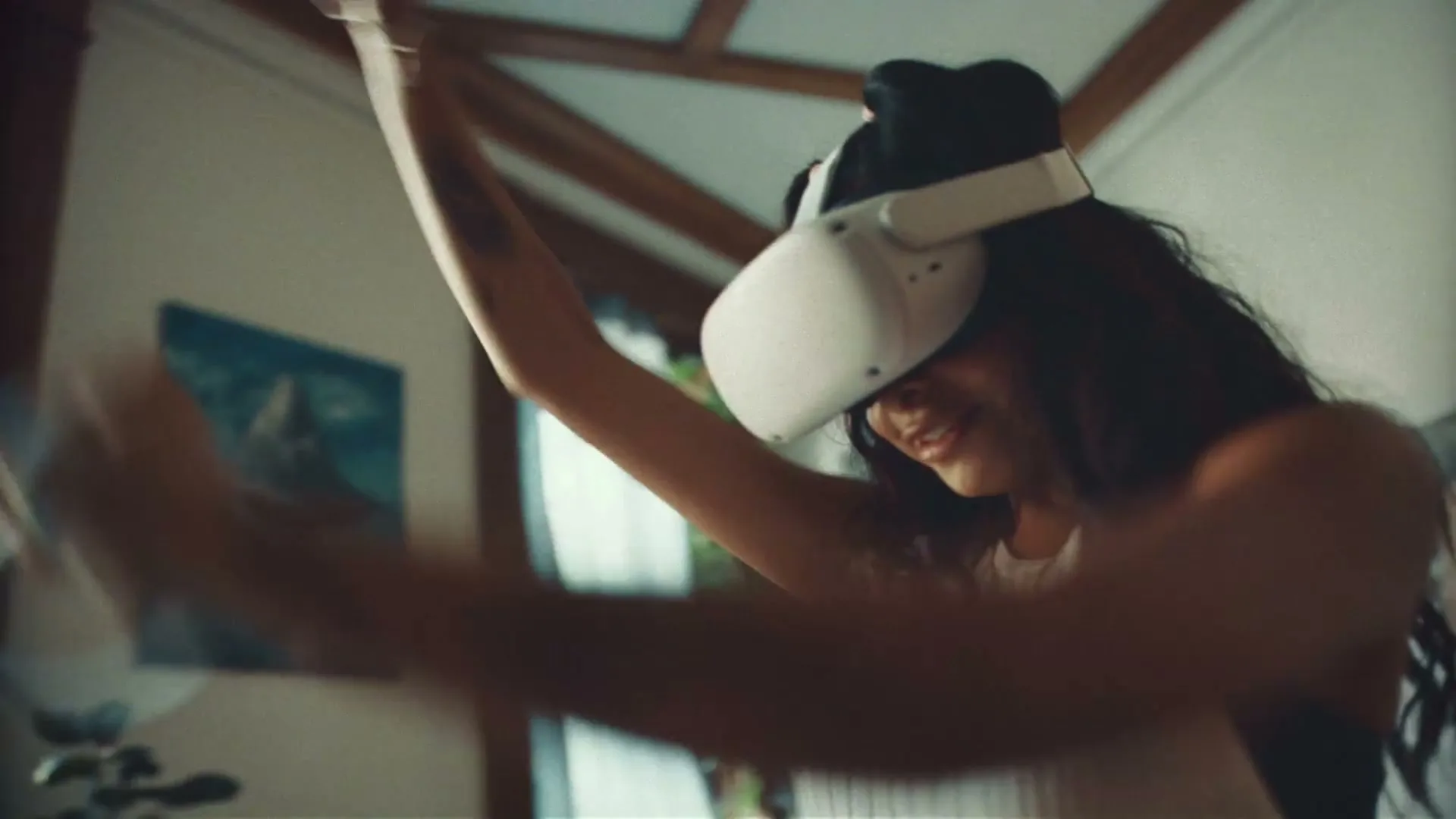 A young woman with long dark hair wears a white VR headset and a white ribbed tank top, smiling and moving her arms in a sunlit room.