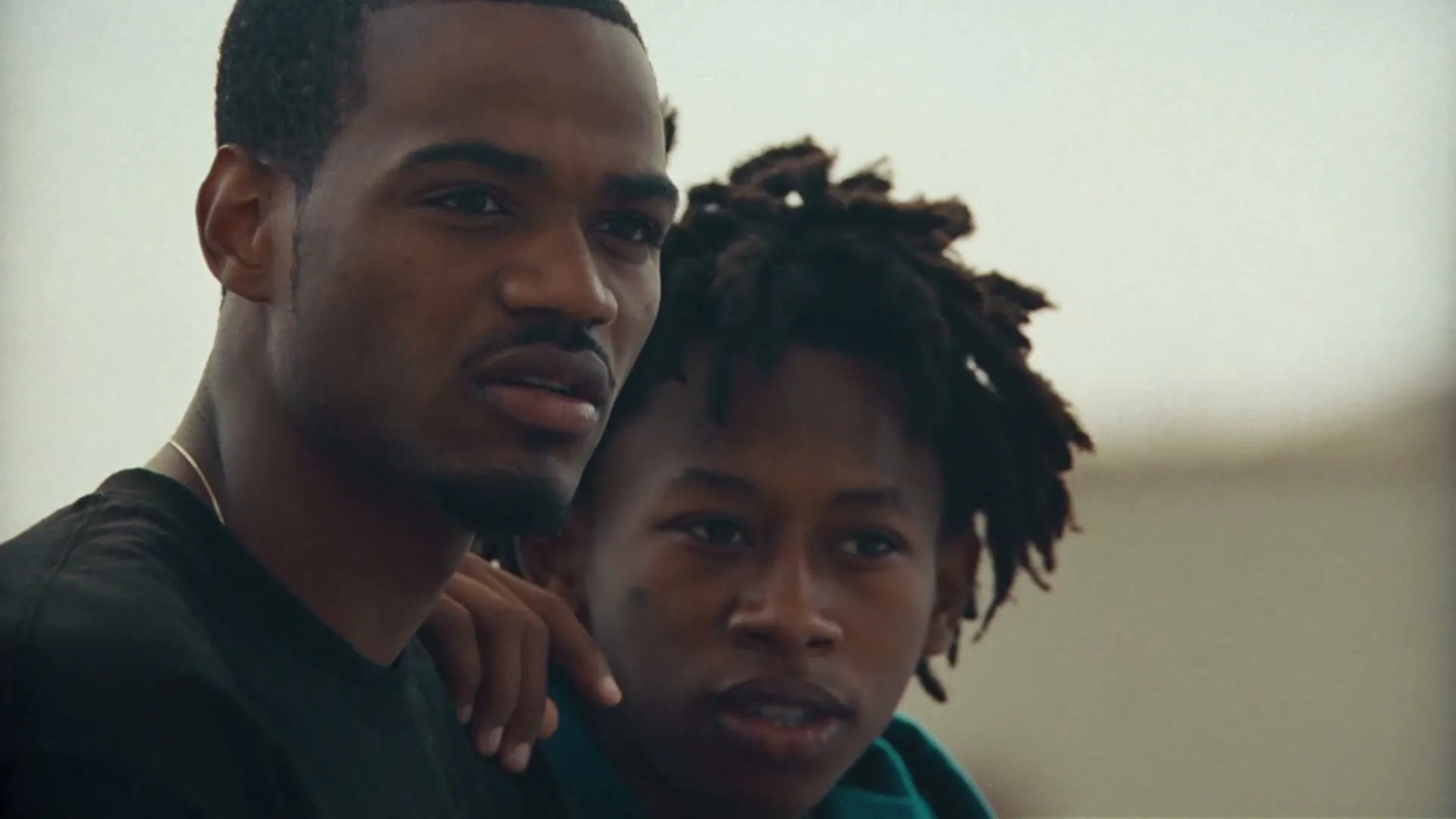 Two young Black men, one older with a hand on the younger's shoulder, look off-camera with serious expressions outdoors.