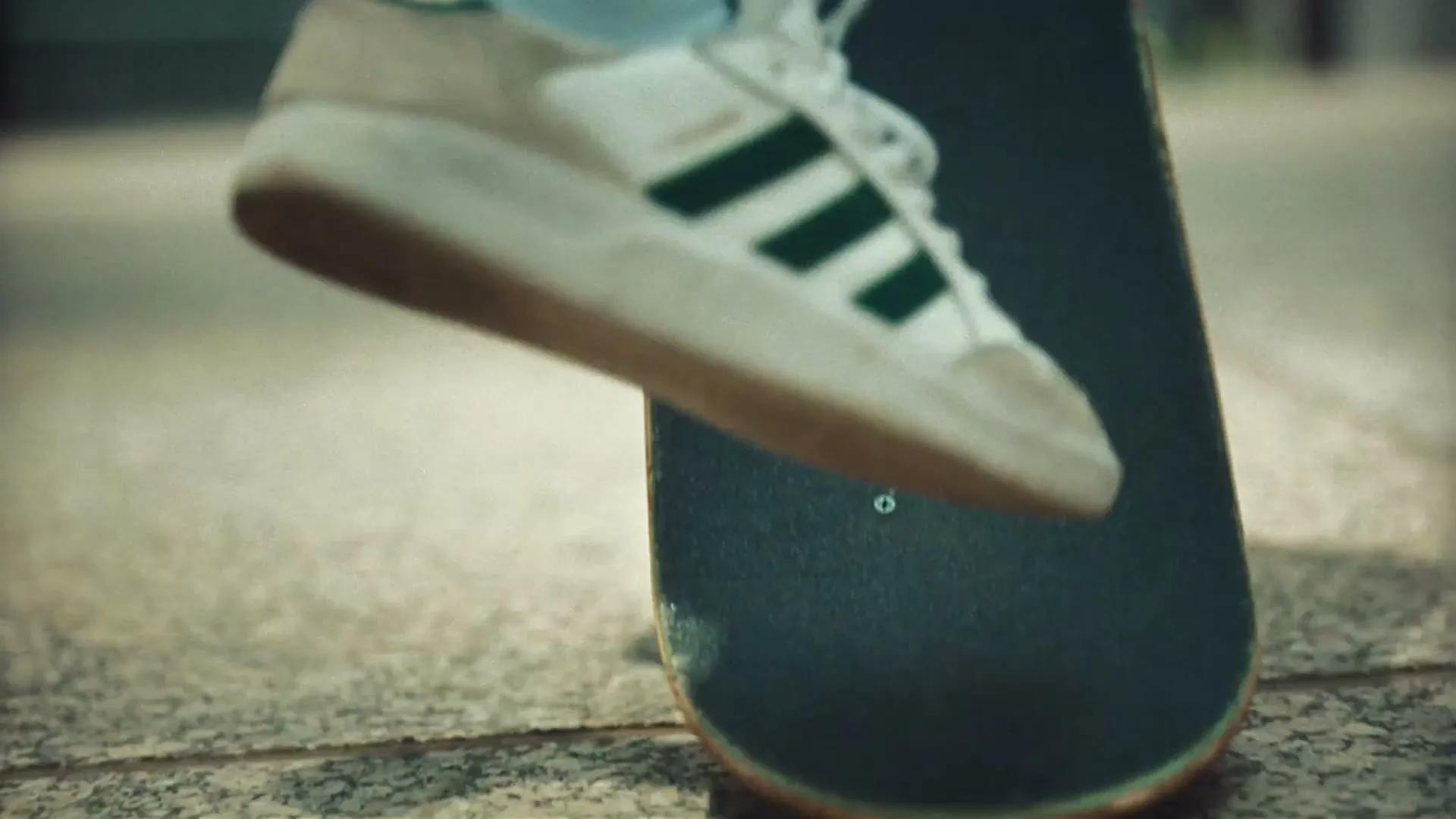 A person's feet in white and green sneakers are on a skateboard, which then rolls away on a paved surface.