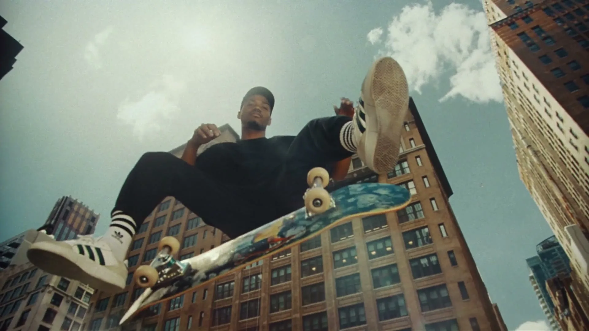 A young Black man in a black t-shirt, black pants, and white sneakers with black stripes, performs a skateboard trick in a city street.