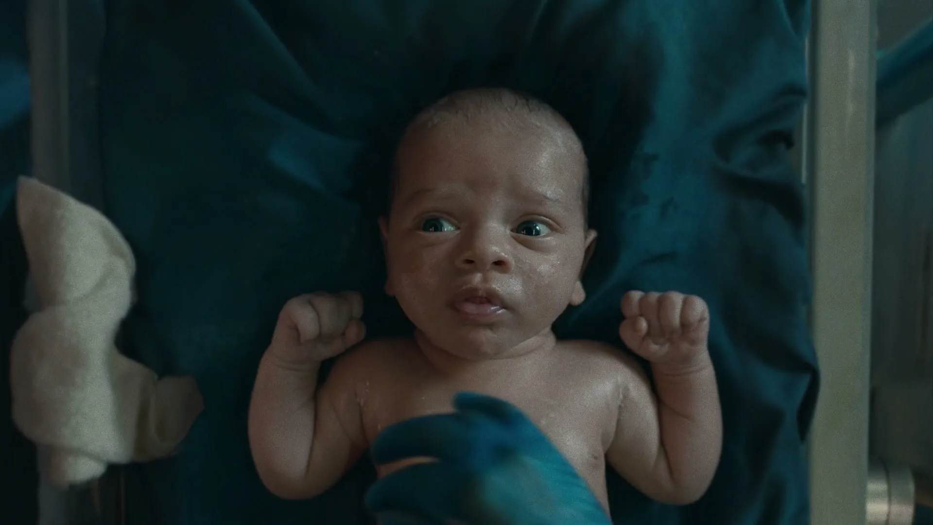 A newborn baby with dark skin and wet hair lies on a blue cloth, looking around as medical staff attend to it in a hospital.