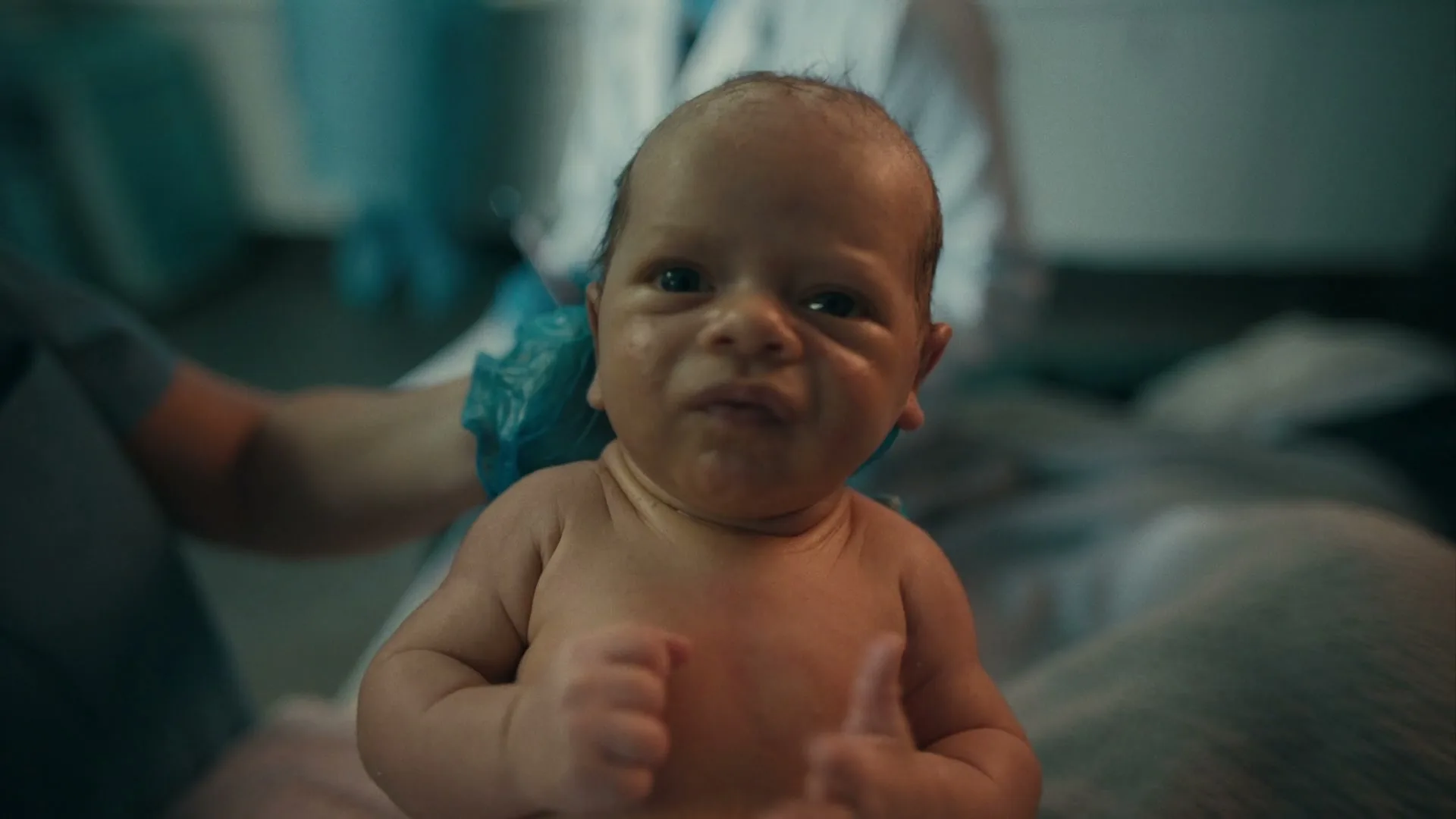 A newborn baby with dark hair and light skin is held by an adult, looking directly at the camera with a scrunched face in a hospital room.