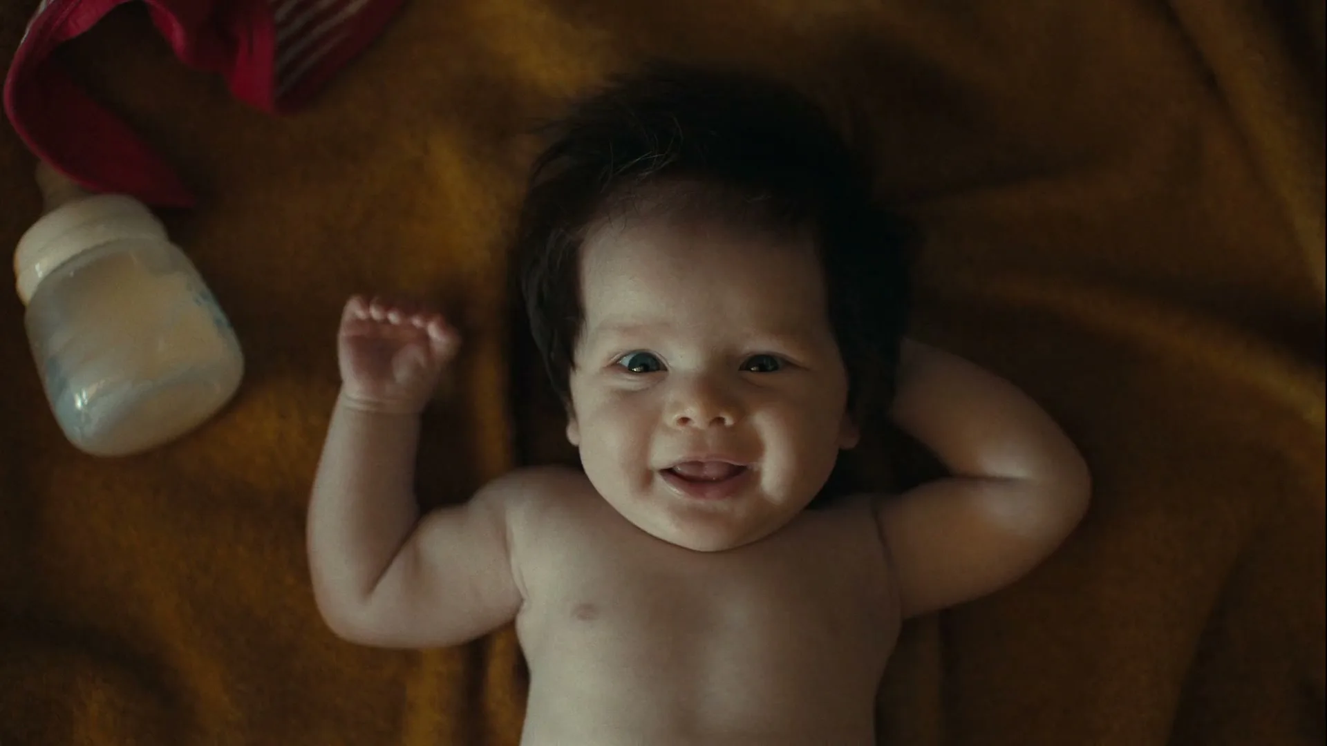 A baby with dark hair lies on a mustard yellow blanket, looking up at the camera and smiling, with a baby bottle nearby.