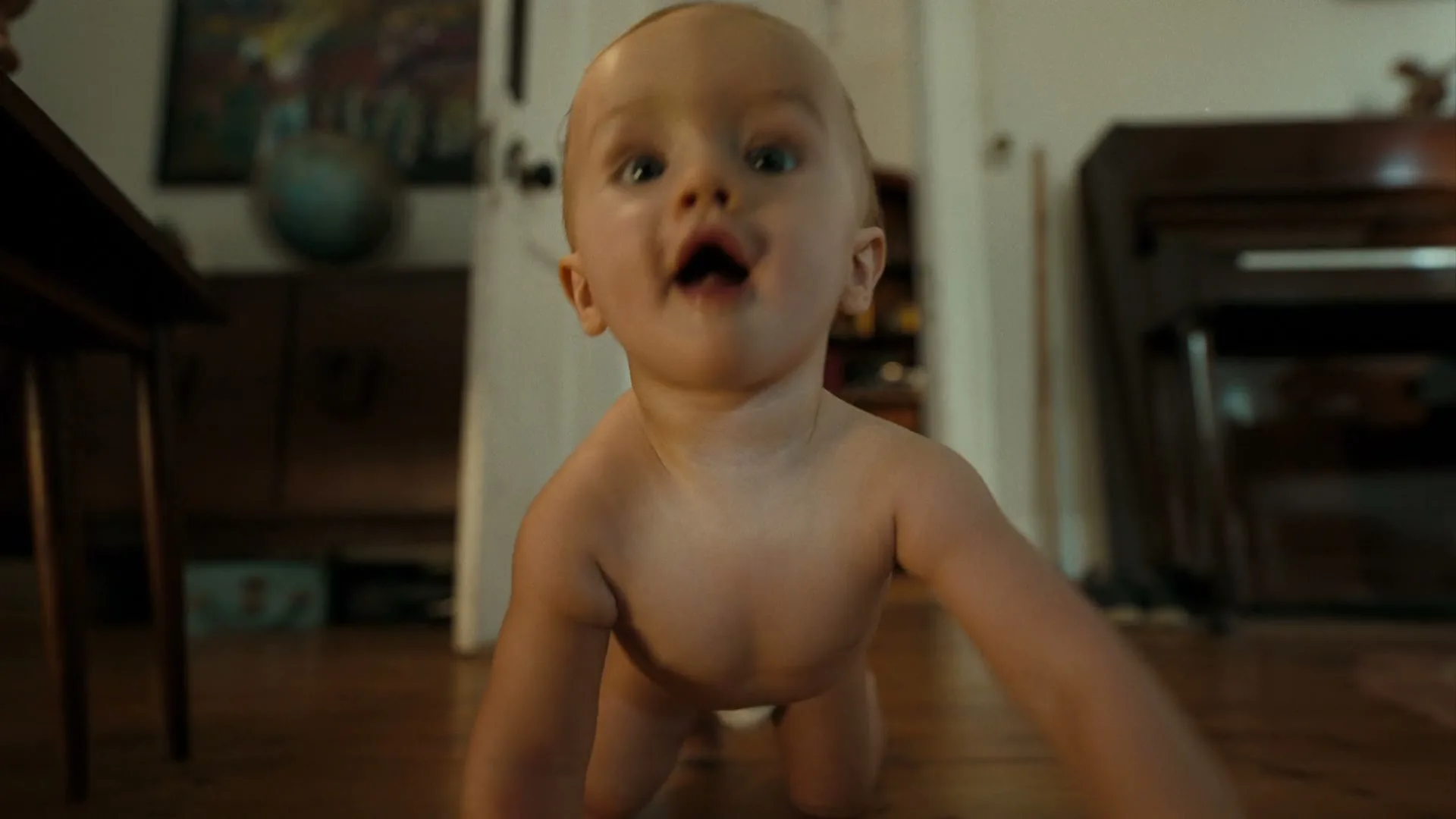 A baby with light skin and hair crawls on a wooden floor towards the camera in a dimly lit room.